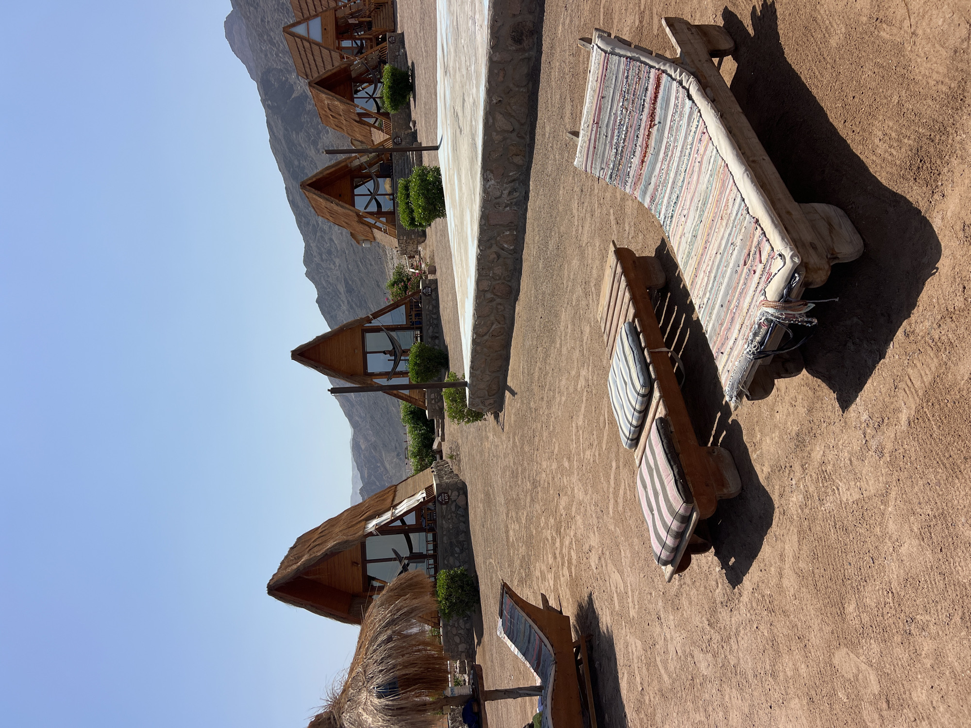 A view of a resort in the city of Nuweiba, Egypt, shows a row of triangular cabins with thatched roofs nestled against a backdrop of arid, mountainous terrain under a clear blue sky. In the foreground, several rustic wooden lounge chairs with striped cushions are scattered across the sandy ground. To the left, a thatched umbrella shades one of the lounge chairs. The cabins, constructed with wood and stone foundations, feature large glass windows that offer glimpses into hammocks strung inside. The overall scene depicts a tranquil, desert-like setting designed for relaxation.