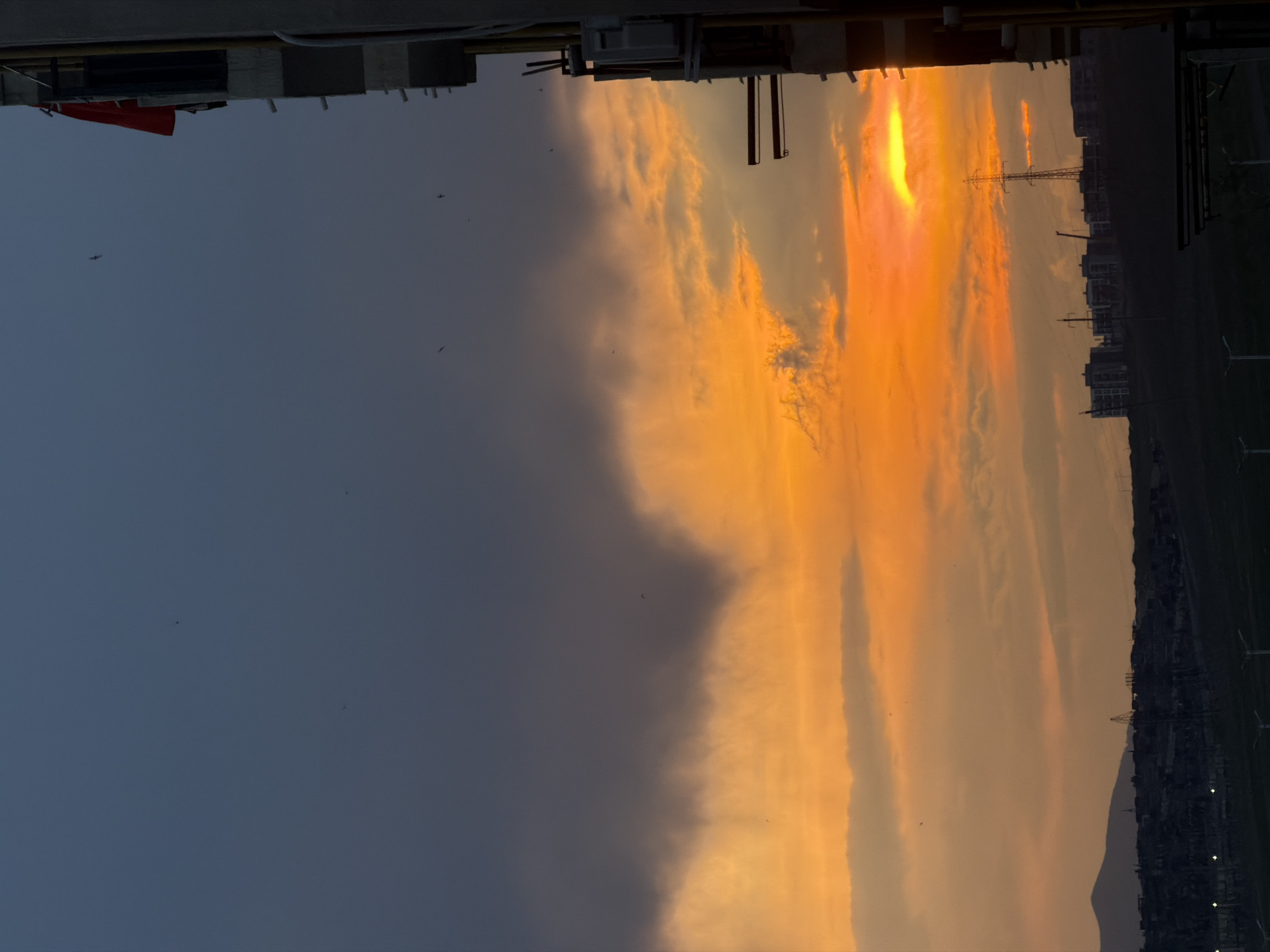 The image displays a dramatic sunset or sunrise over a vast landscape, with the sky dominating the frame. The right side of the sky is illuminated by intense orange, yellow, and fiery red hues, indicating the sun's position near the horizon. These vibrant colors cast a warm glow on scattered clouds, creating a striking contrast with the darker, cooler blue-grey clouds and sky on the left side of the frame. Several small, dark specks, likely birds, are visible against the brighter sections of the sky.
On the horizon, the landscape and urban elements are rendered as dark silhouettes against the colorful sky. A distant city or town skyline stretches across the lower portion of the image, featuring various buildings, tall power line towers, and multiple wind turbines. A faint mountain or hill range is also visible in silhouette behind the urban sprawl.
In the immediate foreground, along the top edge of the image (assuming a corrected landscape orientation where the horizon is at the bottom), parts of a building or structure are visible. This includes what appears to be a balcony railing or rooftop edge, and a small, partially obscured red flag or banner hanging downwards. The photograph was captured from an elevated vantage point, possibly from a building in Tbilisi, Georgia, looking out over the area during twilight. The overall scene evokes a sense of natural beauty and tranquility. No text is visible in the image.