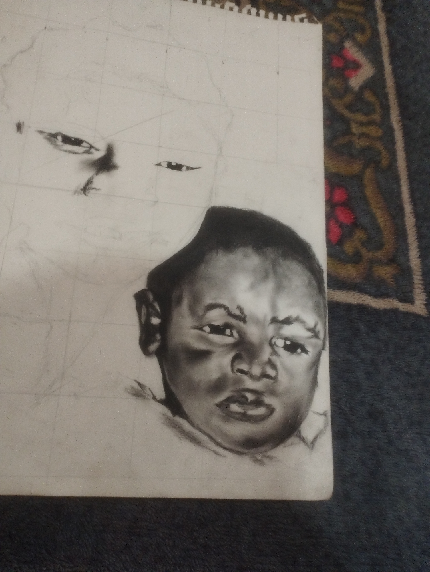 The image shows a charcoal drawing of a baby's face, partially rendered. The baby's face is the most detailed element, with shading and definition around the eyes, nose, and mouth. The drawing is on a white sheet of paper, which has faint grid lines visible. In the background, there are faint pencil sketches of what appears to be another face or head, less detailed than the main subject. The paper is positioned on a dark, textured surface, possibly a rug or carpet with a patterned border visible on the right side. The drawing is incomplete, with a significant portion of the paper remaining blank or lightly sketched. There is no identifiable location in the city of Jos, Nigeria, visible in the image.