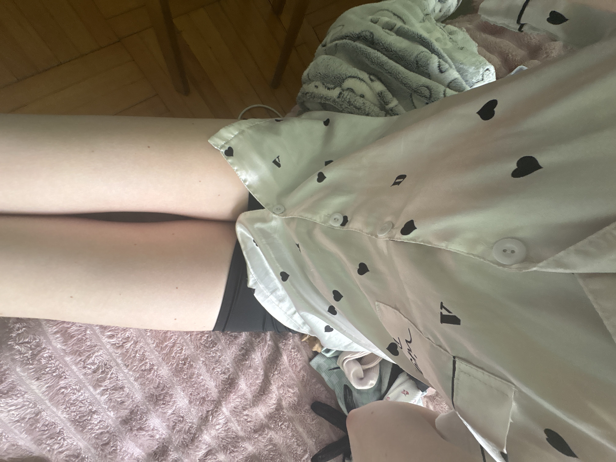 The image shows a person in a white top with black heart patterns and black bottoms, lying on a pink blanket. The top is buttoned and has a pocket on the right side. The person's legs are visible on the left side of the image. There is a fuzzy blanket behind the person. The location is not identifiable from the image.