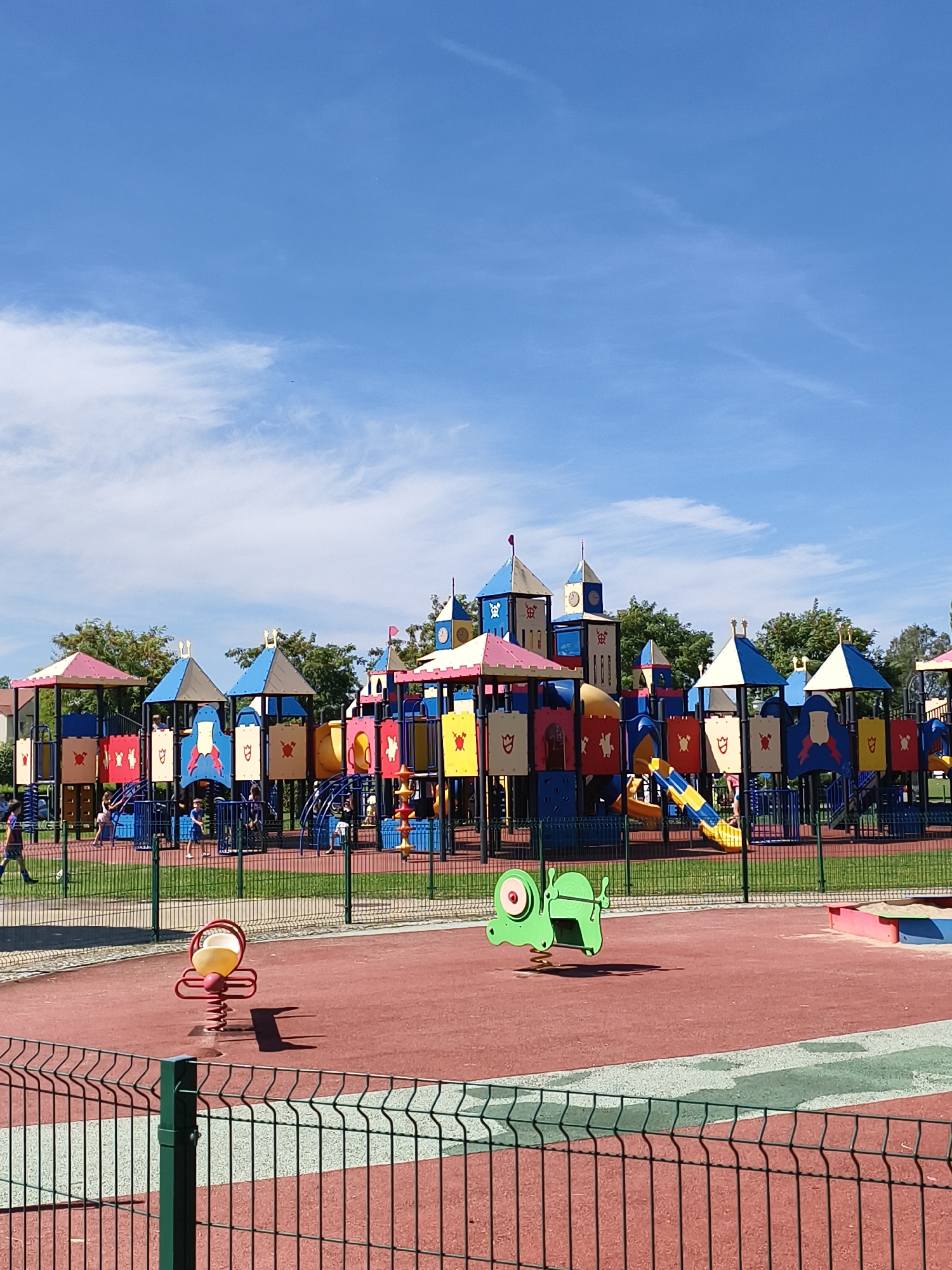 The image shows a vibrant, colorful playground designed to resemble a medieval castle. It features multiple towers, slides, and climbing structures, predominantly in blue, yellow, red, and pink. The playground surface is a red rubberized material with some green surfacing visible. In the foreground, there are two spring riders: one is a yellow egg-shaped seat on a red spring, and the other is a green frog-shaped rider. A black metal fence encloses the playground. Beyond the playground, a line of trees forms a backdrop, and the sky is a clear blue with scattered white clouds. The image appears to be taken on a sunny day. The location is identified as Tczew, Poland.