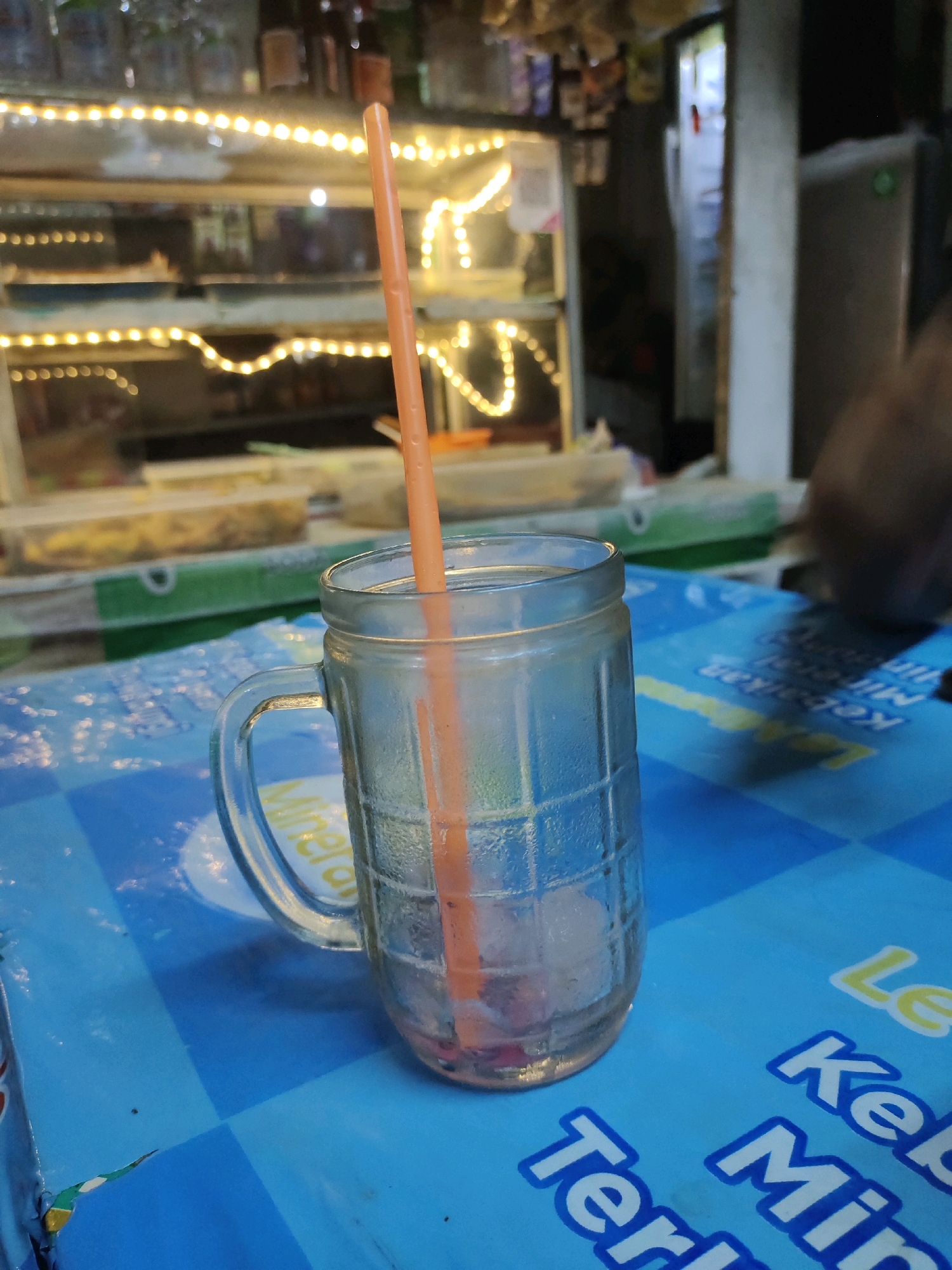 An orange plastic straw rests inside a clear, ribbed glass mug, which sits on a blue and white checkered surface, possibly a table or placemat, in what appears to be an outdoor food stall or small shop. The background is blurred but shows shelves filled with items, some illuminated by strings of warm-toned lights, suggesting it could be evening. A refrigerator unit is partially visible on the right. The overall scene is simple and focuses on the glass and straw, with a hint of a commercial setting. The image is likely taken in Lengkong Kulon, Indonesia, given the context of similar images.