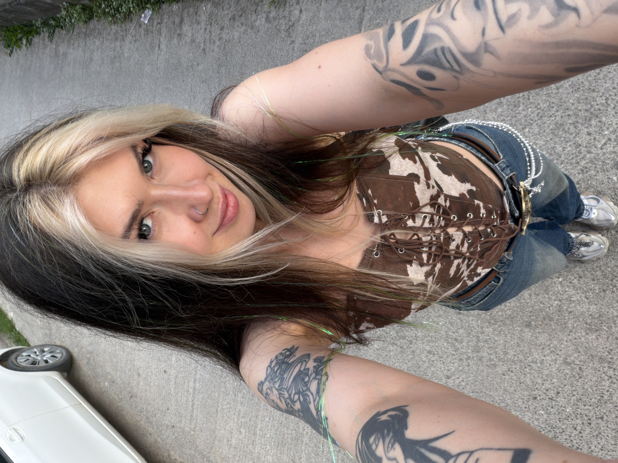 A woman is taking a selfie at an upward angle. She has fair skin, green eyes, a nose ring, and dark hair with a blonde streak. She is wearing a brown and white corset-style top with lacing, blue jeans, and a jeweled belt. She has multiple tattoos on her arms. The background shows a gray asphalt surface, a white car, and some greenery. The location is not discernible from this image.