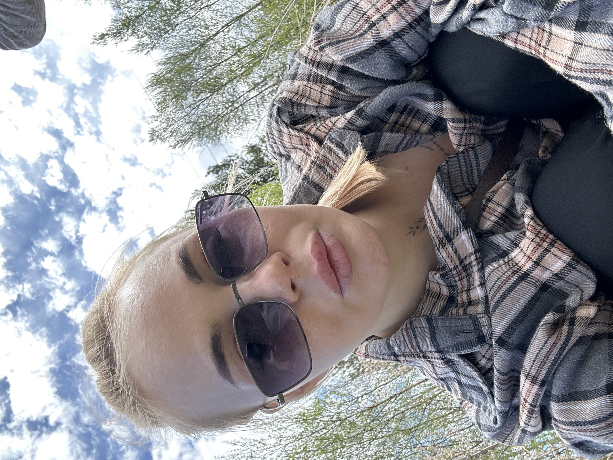 A woman with fair skin and blond hair is wearing sunglasses and a plaid shirt. She is outdoors. The sky is visible in the background. The location is the city of Vysokiy, Russia.