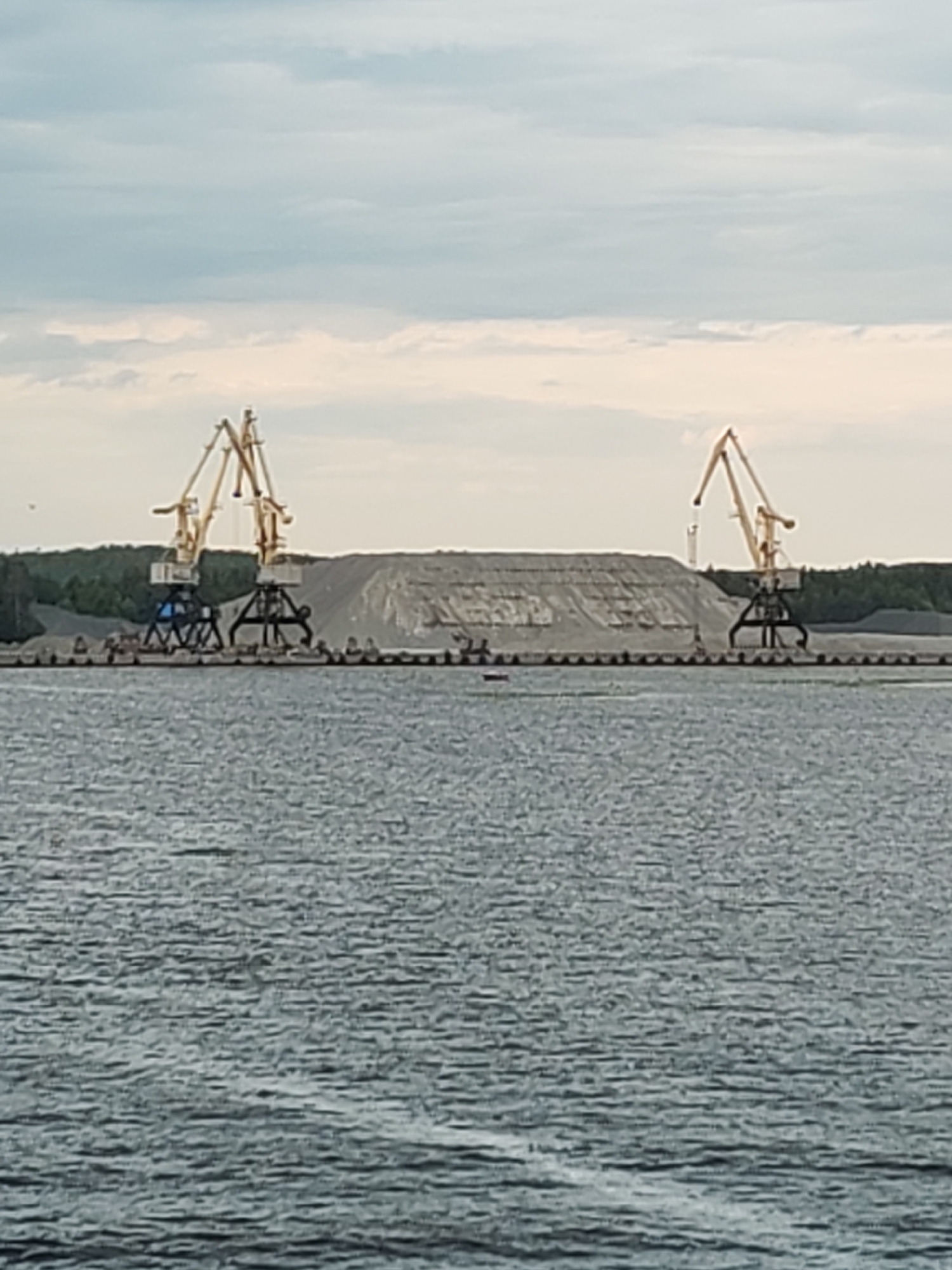 The image depicts a waterfront industrial scene in Shalsky, Russia. Two large yellow cranes stand on a dock, overlooking a vast expanse of choppy gray water. Behind the cranes, a massive pile of light-colored aggregate, likely sand or gravel, is visible. The aggregate mountain is flanked by a dense green forest and a grayish-blue sky with scattered clouds. Several smaller objects, possibly buoys or debris, are scattered on the water's surface. The overall impression is one of industrial activity at a port or quarry.