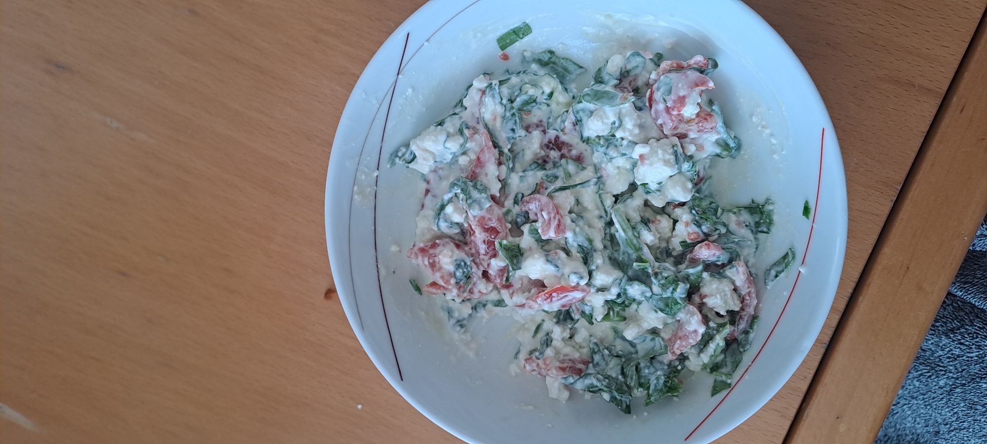 A close-up, overhead shot displays a white bowl filled with a creamy mixture of chopped tomatoes, spinach, and what appears to be feta cheese. The ingredients are finely diced and coated in a white dressing, creating a textured salad. The bowl has a subtle red rim with a thin, dark red line. It rests on a light brown wooden surface, likely a table, with a section of a grey textured fabric visible in the bottom left corner of the frame. The lighting is natural, coming from above and slightly to the left, casting soft shadows. This scene does not contain any people or specific landmarks that would identify its location in Castlebar, Ireland.