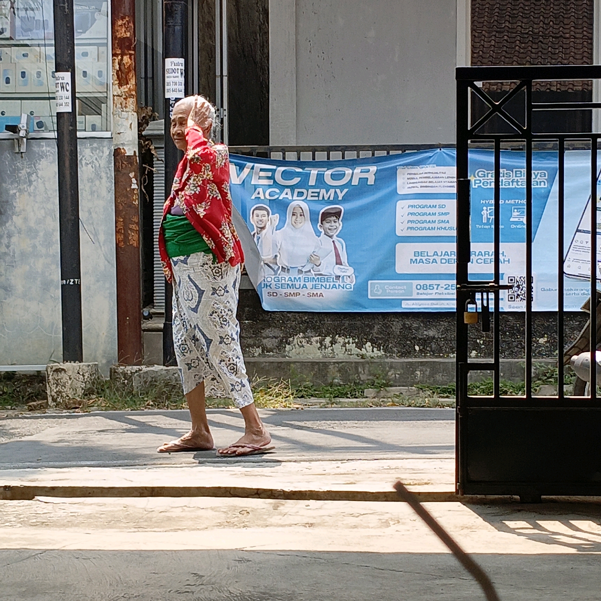The image depicts an elderly woman walking on a paved street in Salatiga, Indonesia. She is wearing a red, floral patterned blouse, a green sash around her waist, and a light-colored patterned skirt. Her head is covered with a patterned cloth, and she has her hand raised to her head. Behind her, a blue banner for "VECTOR ACADEMY" is visible, advertising various programs and offering free registration. The banner also features cartoonish illustrations of students. To the right of the banner, a black metal gate with a yellow padlock is visible, securing an entrance. The environment appears to be a street with some vegetation and buildings in the background. The lighting suggests it is a sunny day, casting shadows on the ground.