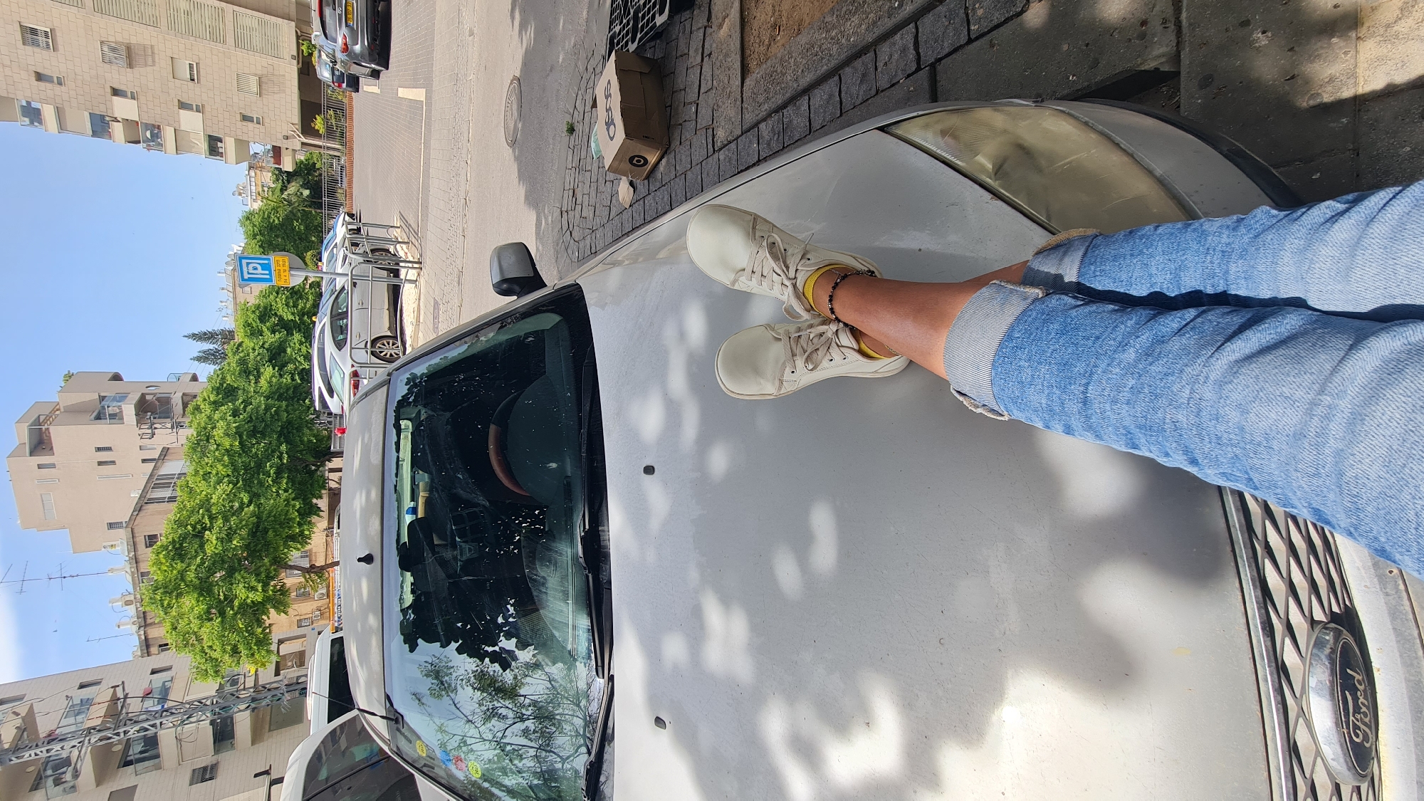 The image is taken in Rishon LeZion, Israel. The view is from the hood of a silver Ford car. A person wearing blue jeans and cream-colored sneakers rests their legs on the car's hood. A brown ASOS box sits atop a bricked curb, along with another object. The surrounding environment includes buildings and trees that are typical of a city setting. A parking sign is also visible across the street.