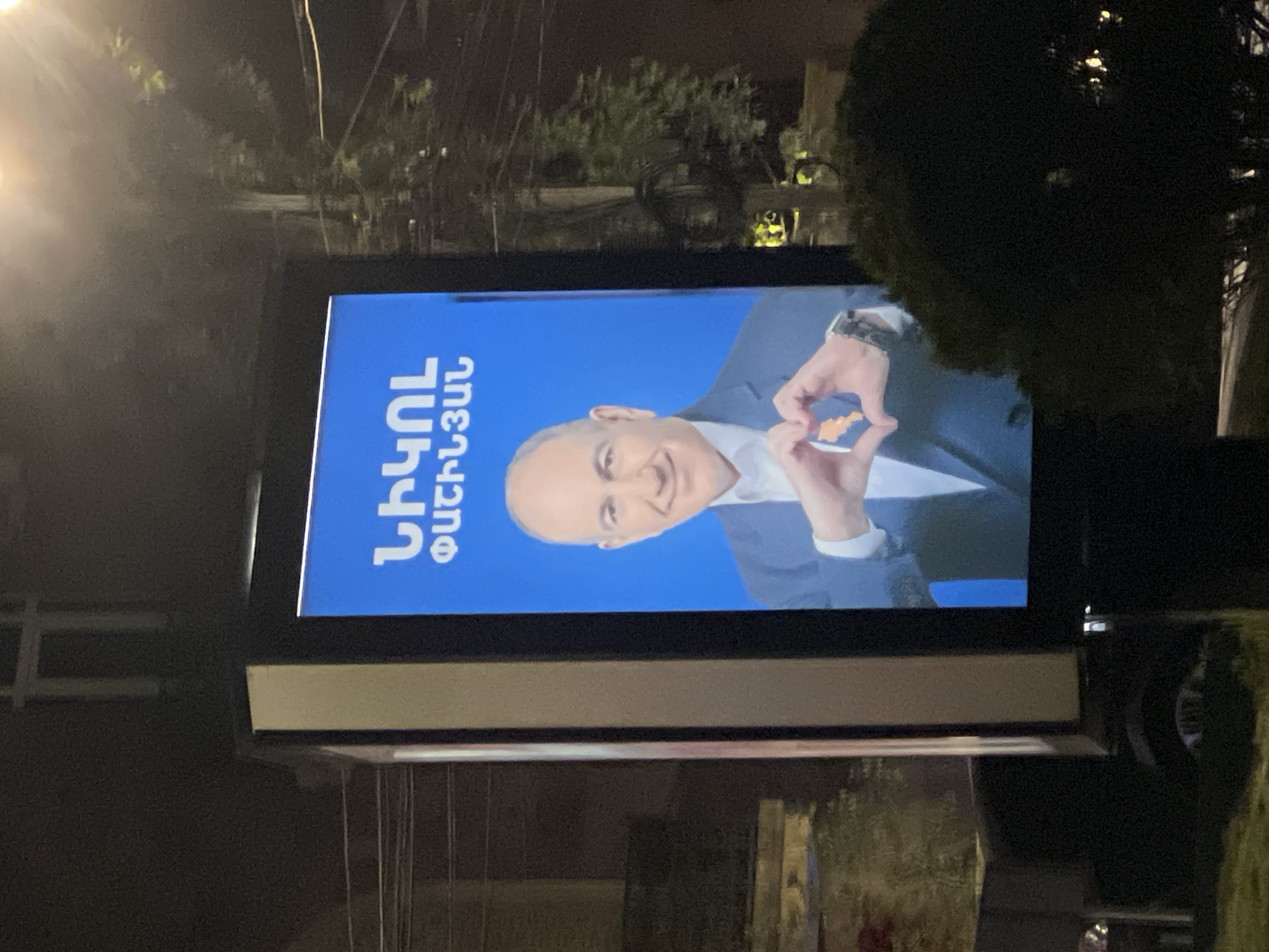 The image shows a billboard in Yerevan, Armenia, featuring a portrait of Armenian Prime Minister Nikol Pashinyan. He is smiling and making a heart shape with his hands. The billboard is lit and appears to be photographed at night. The background includes trees, street lights, and glimpses of buildings. There are also visible wires above.