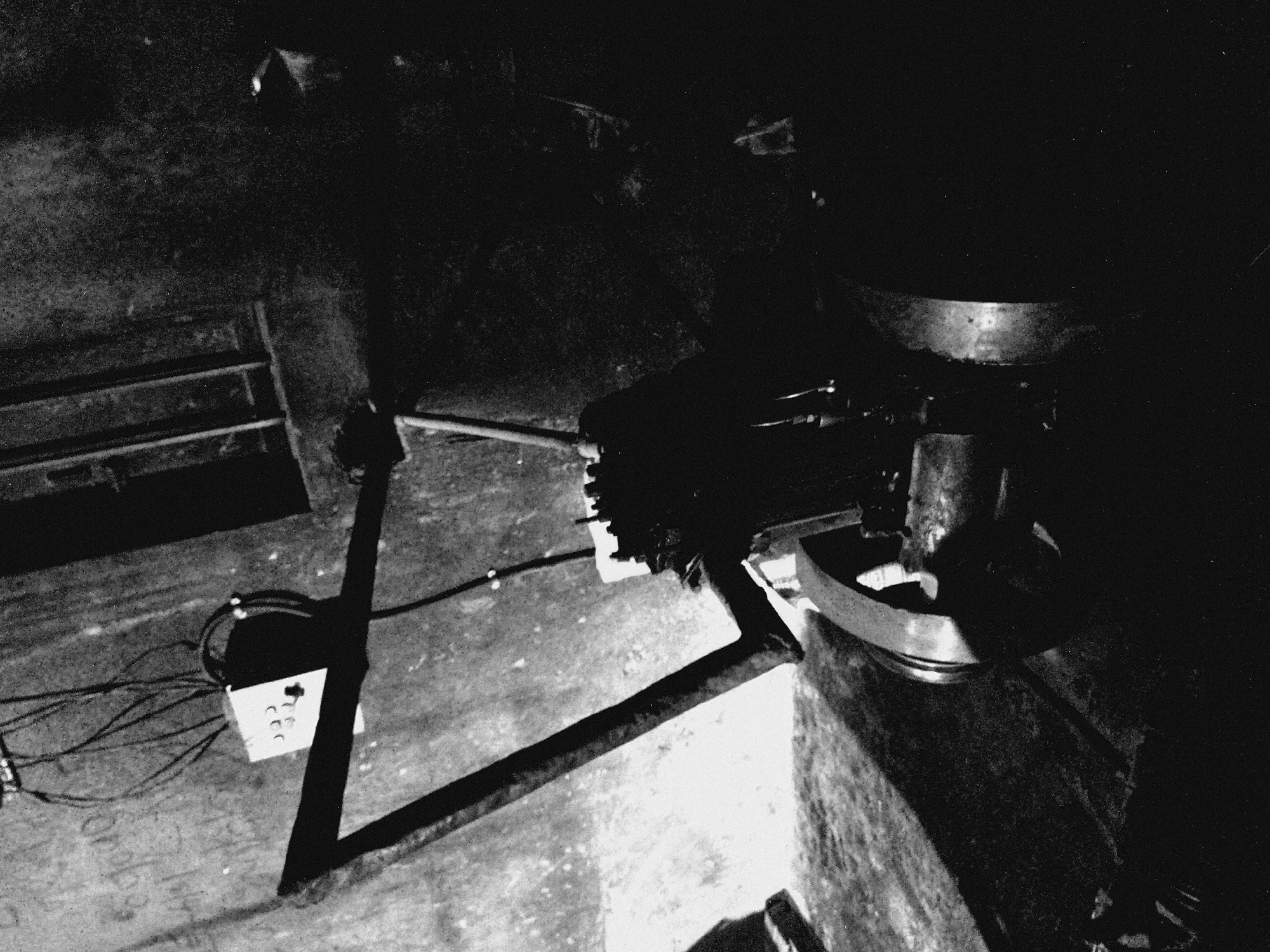 This is a grainy, high-contrast, black and white photograph of an industrial or utility setting, characterized by deep shadows and stark highlights. No people are visible.
The scene features various pieces of mechanical or structural equipment. In the foreground on the left, an electrical junction box, white with several circular connectors, is visible on a concrete floor, surrounded by multiple dark cables. Faint, indistinct characters, possibly numbers like "0003," are faintly visible on the concrete near the box.
Several dark metal beams or supports crisscross the floor, forming part of a frame or structure. On the right side of the image, a large, rounded metal object, possibly a vat, basin, or part of a machine, is prominent. Surrounding this object are other dark, complex mechanical components and machinery, largely obscured by shadow.
The overall environment suggests an indoor space, such as a workshop, factory floor, or basement, due to the concrete floor and lack of natural light. The lighting is very dim, contributing to the high contrast and the overall somber, utilitarian atmosphere. There is no specific activity taking place; the image captures a static arrangement of industrial elements. No discernible text beyond the faint floor markings is clearly visible.