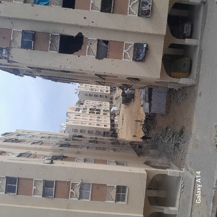 The image depicts a severely damaged urban residential area within Khan Yunis, Palestinian Territory, viewed from an elevated vantage point. Two multi-story, beige apartment buildings frame the foreground, displaying significant damage with shattered windows, missing frames, and large dark apertures indicative of structural impact, particularly on the right building. The ground floors feature arched supports, with a green tarp-covered object and a white car visible under the right structure. Between these buildings lies a vast, disturbed dirt expanse strewn with substantial rubble and debris. A makeshift tarp shelter is situated in the midground. Three individuals are observed within the open area: one person walks near the base of the left foreground building, and two others are positioned further back amidst the debris near the shelter. Numerous similarly damaged multi-story buildings extend into the background, many with rooftop water tanks. Overhead utility lines span the scene beneath an overcast sky. A paved area occupies the immediate foreground.