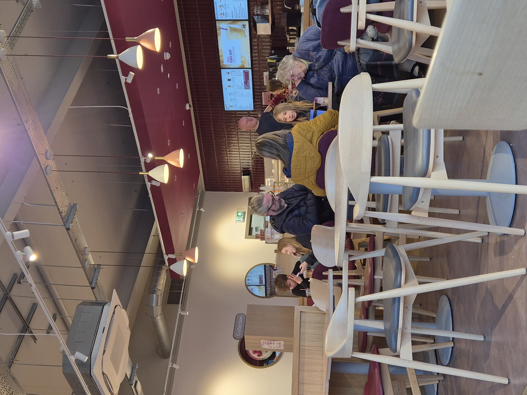 The image depicts the interior of a Costa Coffee shop, likely located in West Suffolk, United Kingdom, as that's where the picture was taken. People are seated at tables, some engaged in conversation while others look at their devices. There are digital menu boards displaying various food and drink options. The interior design features a combination of light-colored wood furniture, white walls, and a burgundy-colored ceiling accent with pendant lights. A "Collection" point sign is visible on the wall, suggesting an area for order pick-up. The ceiling is lined with industrial-style fixtures and ventilation systems.
