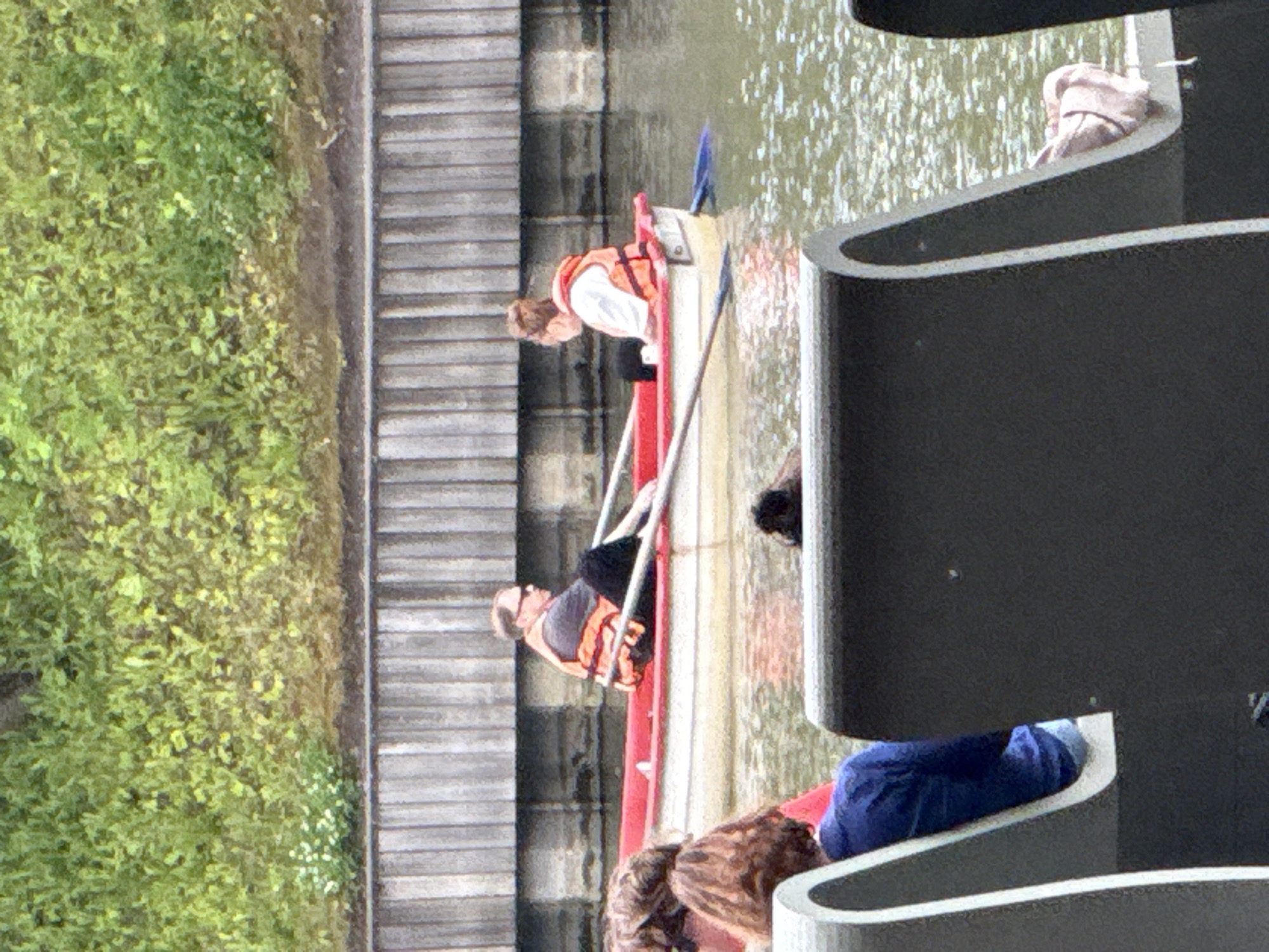 The image shows a view of the Moscow Canal in Russia. In the foreground, two figures are seated on dark grey benches, facing away from the camera. One figure appears to be wearing a blue top. In the middle ground, a red and white rowboat is on the water. Two people are in the rowboat, both wearing orange life vests. One person is rowing the boat with two oars. The background consists of a wooden wall and dense green foliage.
