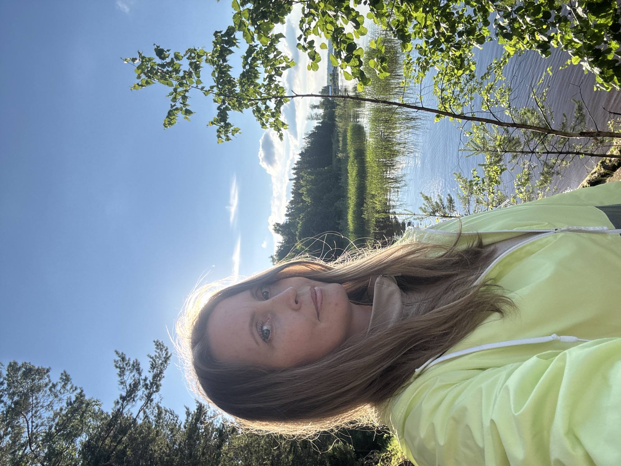 A medium shot captures a woman in her late 40s or early 50s, standing outdoors by a body of water, likely a lake or river, under a clear blue sky with scattered clouds. The woman has long, light brown hair that is partially backlit by the sun, creating a luminous effect around her head. She is wearing a light yellow hooded jacket over a beige turtleneck. Her expression is calm and she is looking slightly to her right, towards the camera.
The background features a tranquil natural scene. To her left, there are tall pine trees. To her right and behind her, there is a wide expanse of water with reedy vegetation growing along the edge. Across the water, a dense forest line with varied green trees stretches towards the horizon. The overall lighting suggests it is late afternoon or early evening, with the sun casting a warm glow.
The image is a selfie, indicated by the perspective and the woman's direct gaze. No specific landmarks or identifying features of Chaikovsky, Russia are visible in the scene; it depicts a typical natural landscape that could be found in many regions with forests and bodies of water.