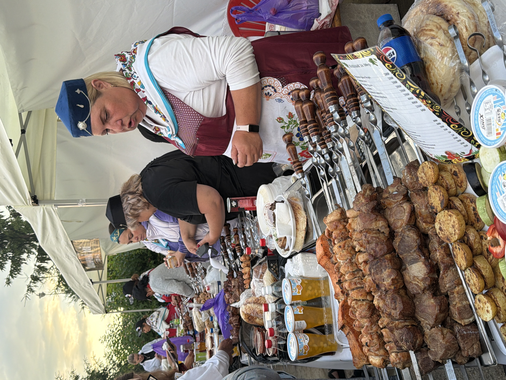 In an outdoor market setting, likely a food stall, a woman wearing a blue cap and a decorative apron stands behind a table laden with various grilled meats and other food items. She appears to be interacting with customers, possibly taking an order or serving food. The table displays rows of skewers filled with cooked meat, as well as grilled vegetables, bread, and drinks in cups. Other people are visible in the background, some browsing the offerings, while others seem to be working at adjacent stalls. The overall atmosphere suggests a busy marketplace during the daytime, possibly with a festive or cultural element given the traditional attire of some individuals. The background shows tents and trees, indicating an outdoor event.