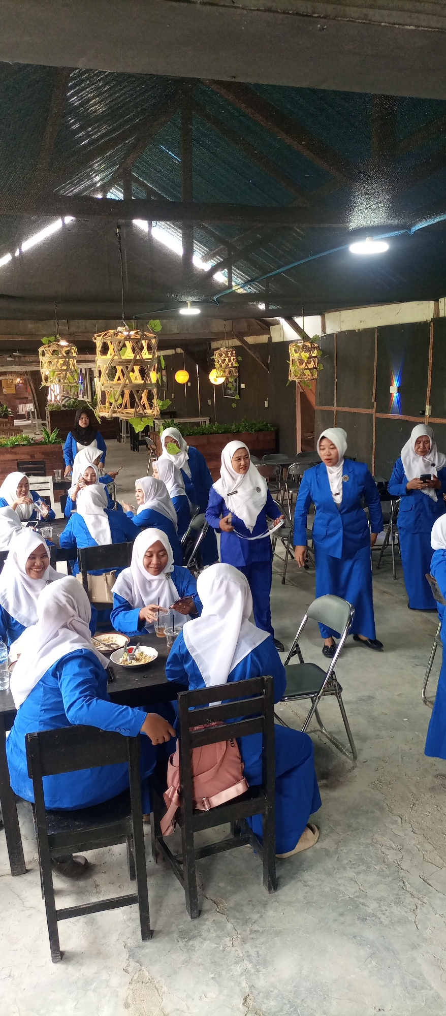 The image depicts a gathering of women wearing blue uniforms and white hijabs inside a rustic, open-air establishment, likely a cafe or restaurant, in Kedewan, Indonesia. The ceiling is constructed of corrugated metal and exposed wooden beams, with decorative bamboo light fixtures hanging down. Several tables are present, with some women seated and others standing.
In the foreground, several women in blue uniforms are seated at a table. One woman in the center is looking at her phone. To her right, another woman is also seated and looking forward. To the left, two other women are seated, one of whom is looking towards the center. A plate of food and drinks are on the table.
Further back, more women in similar attire are either seated or standing, engaged in conversation or observing their surroundings. One woman in the far right is standing and looking at her phone. The overall atmosphere appears casual and social. The floor is concrete. Plants are visible in planters along the walls, suggesting an attempt to add greenery to the space. The lighting is a mix of natural light filtering through gaps in the roof and artificial lights from the fixtures.