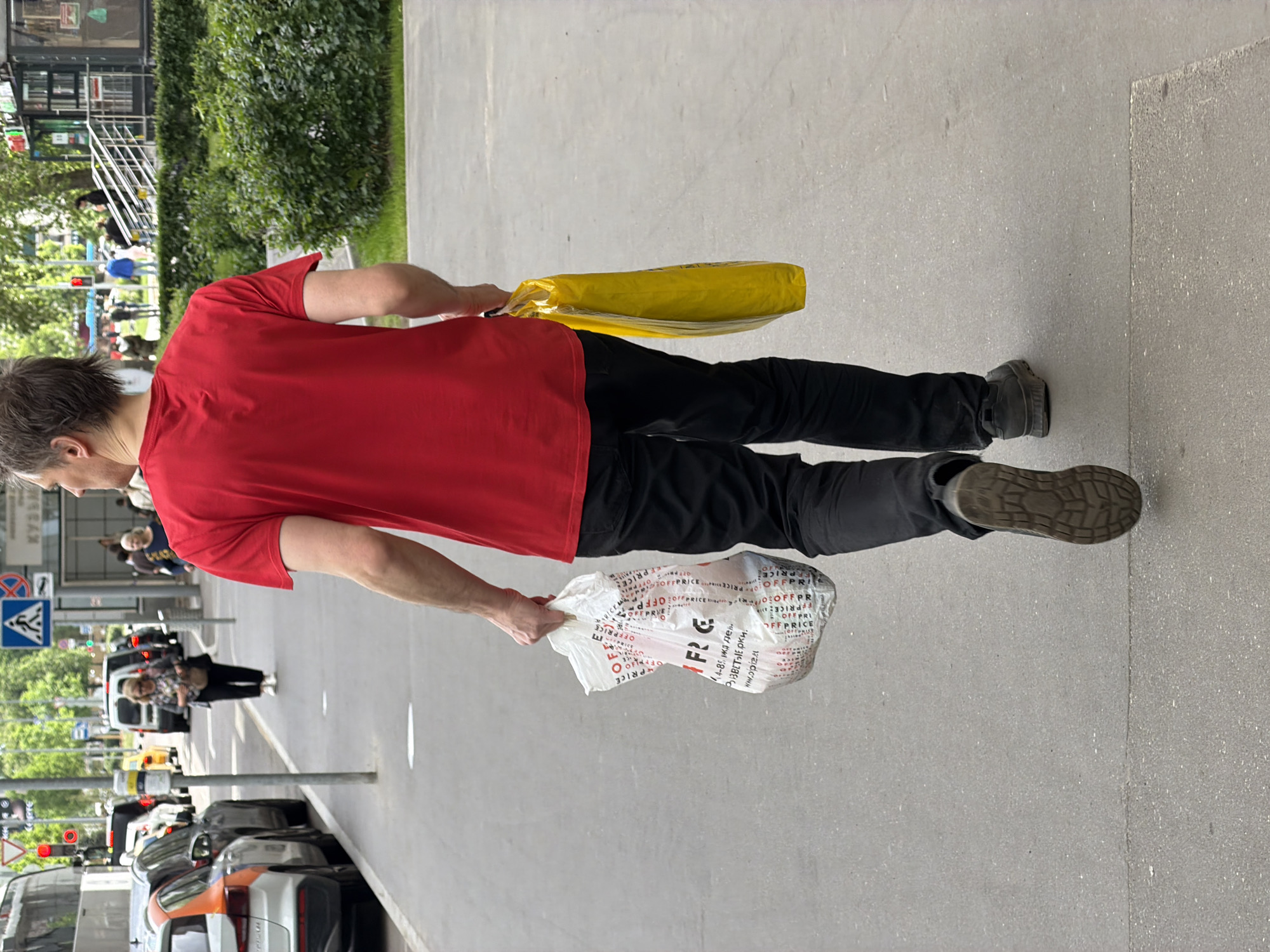 A man is walking on a city street in Unknown, Unknown. He is wearing a red shirt and black pants. He is carrying a yellow package in his right hand and a white plastic bag with writing on it in his left hand. In the background, there are cars, buses, a street crossing sign, and people. There are trees and greenery in the background on the left side of the image. The street is paved and the sidewalk is paved.