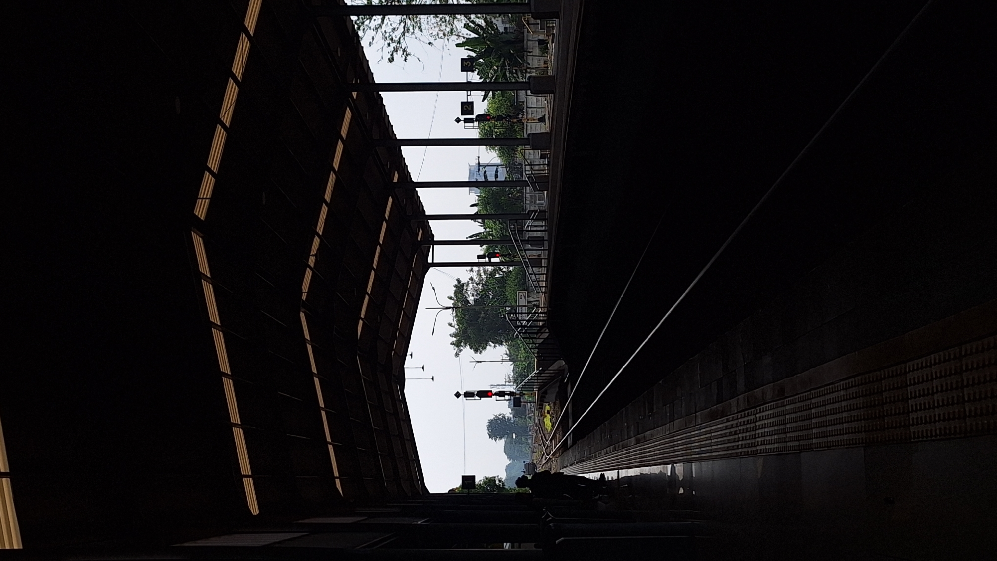 The photograph depicts a railway station in Karawang, Indonesia, taken from an elevated platform. The station is partially covered by a dark, angled roof structure with horizontal slits allowing slivers of light to penetrate. Across the tracks, signals stand, with red lights illuminated. Greenery is visible beyond the railway infrastructure, including trees and other vegetation. A person is barely visible in shadow on the platform. The station platform has a patterned tile surface and a tactile paving strip along the edge. The view extends into the distance, showing structures partially obscured by trees.