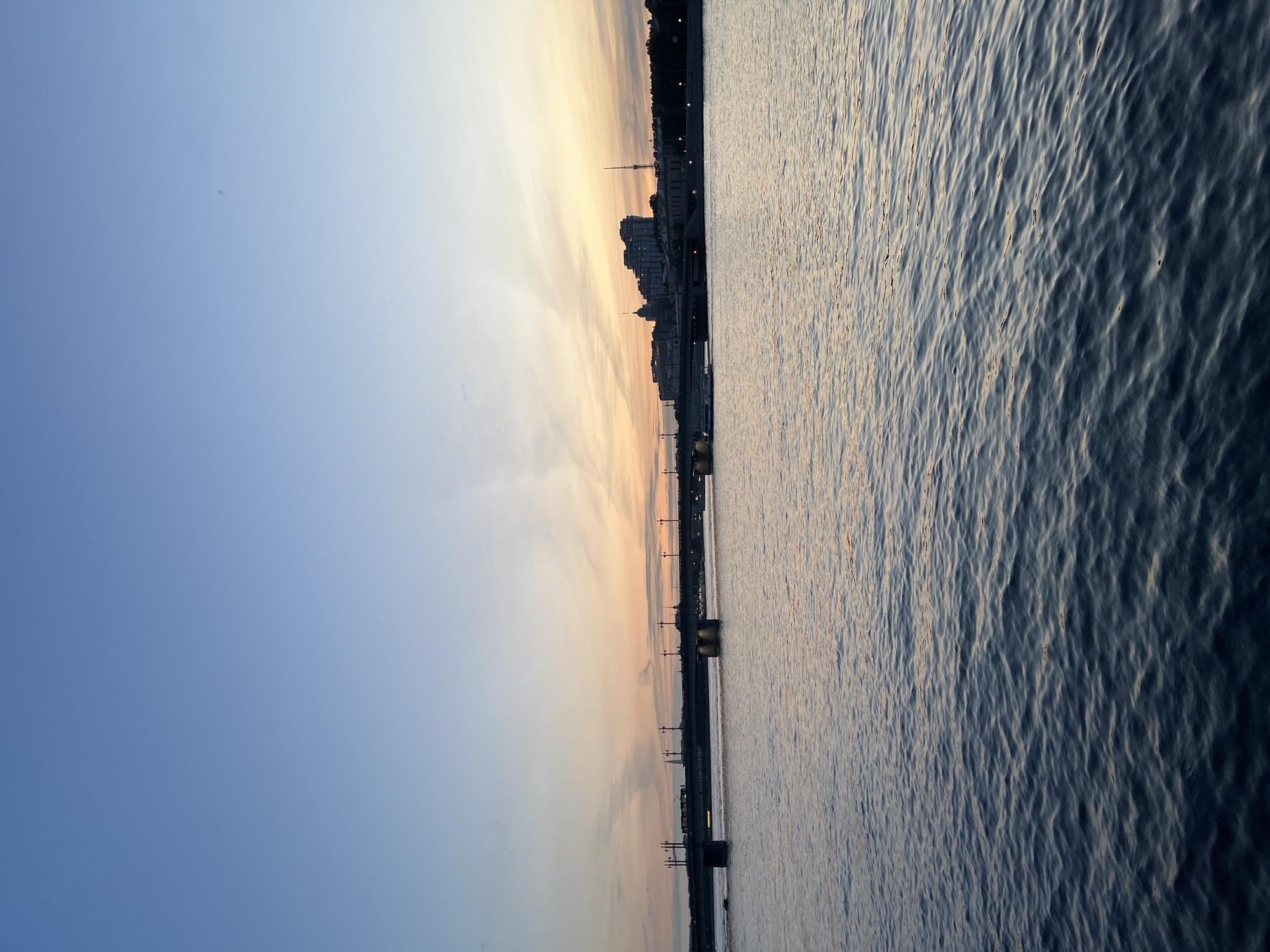 The image shows a wide river with a city skyline in the background. The sky is a gradient of blue and orange, indicating either sunrise or sunset. A bridge spans across the river, with streetlights visible along its length. Buildings of various sizes and architectural styles line the riverbank, with some taller structures appearing to be apartment buildings or offices. The water is calm, with ripples reflecting the colors of the sky. A boat is visible on the river, passing under the bridge. The overall scene is peaceful and serene, capturing a moment of natural beauty in an urban setting.