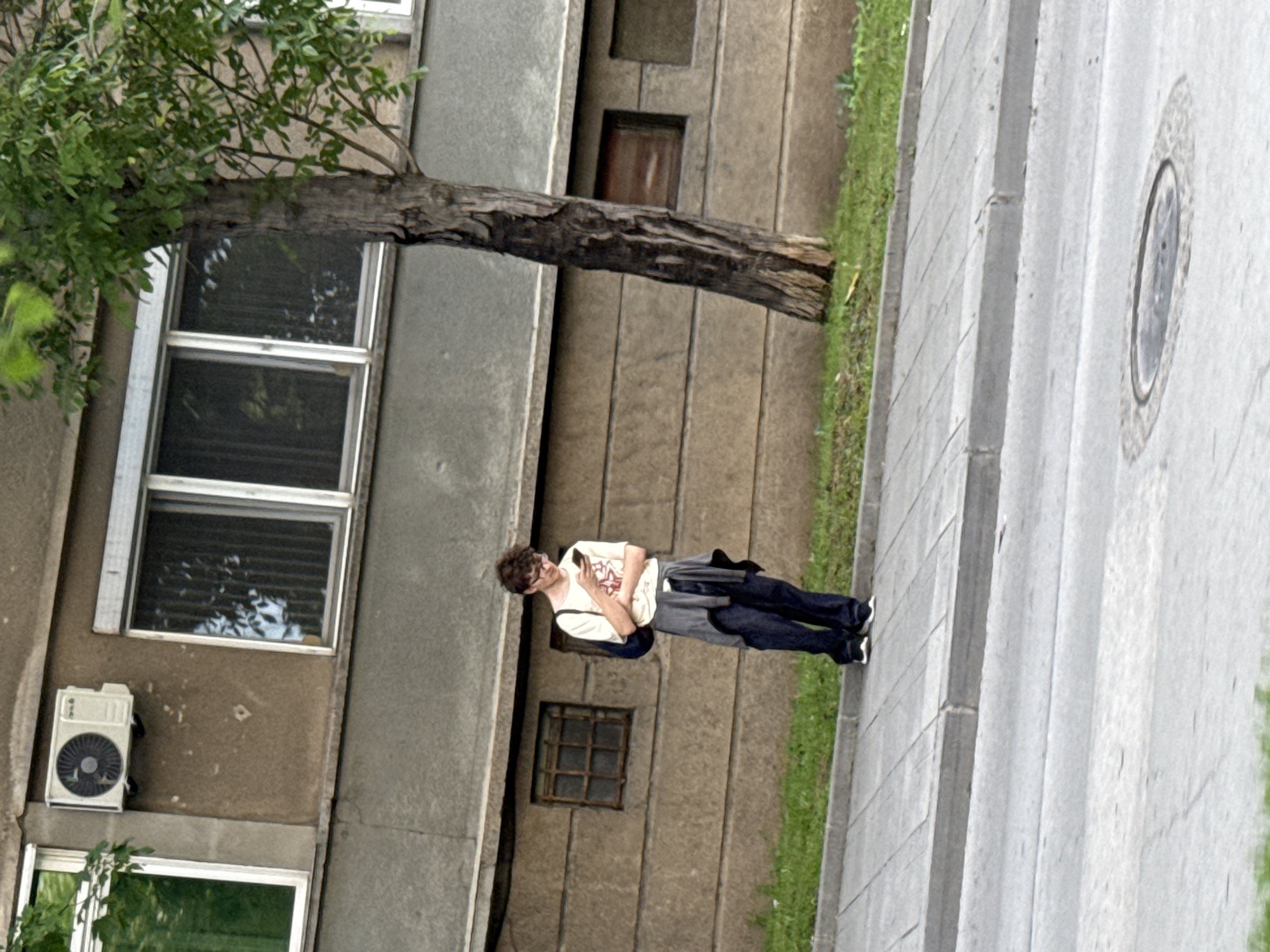 A man stands on a sidewalk next to a concrete building in Pleven, Bulgaria. The building has multiple windows and a tree trunk extending out from it. The man is wearing a light-colored t-shirt, dark pants, and glasses, and he is looking down at a mobile phone in his hands. He has a jacket or sweater slung over his shoulder. The sidewalk is made of concrete slabs. The image is slightly rotated, giving a tilted perspective of the scene.