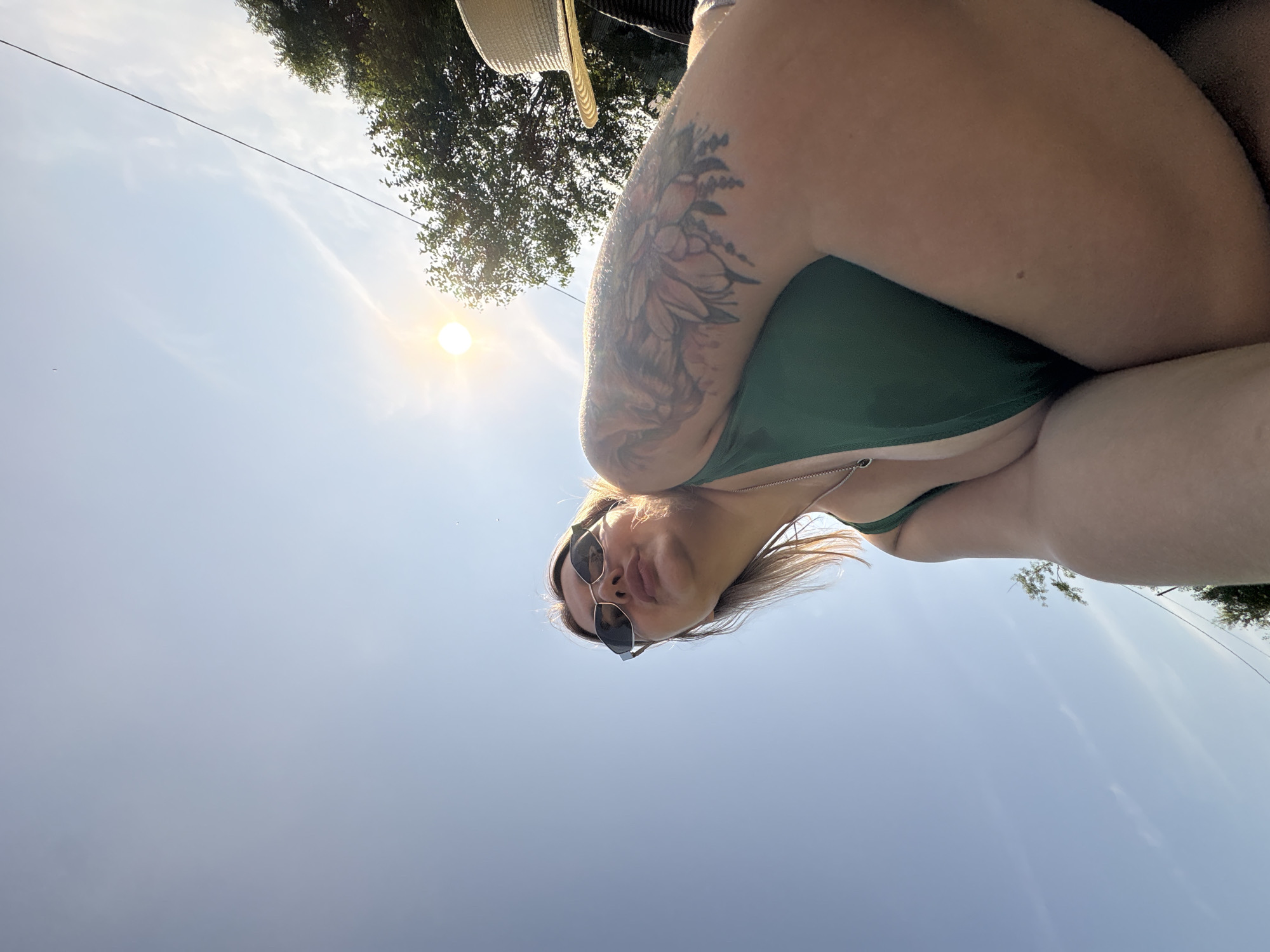 A low-angle, rotated shot shows a person in a green bikini top, wearing sunglasses and a necklace. The person has blonde hair and a tattoo on their upper arm. They are outdoors with a bright sun in the sky, indicating daytime. Trees are visible in the background, and a straw hat is perched at the top of the frame. The scene appears to be a sunny day in Razdolnoye, Russia. The sky is clear with some wispy clouds. The image is taken from a perspective that looks up towards the sky.