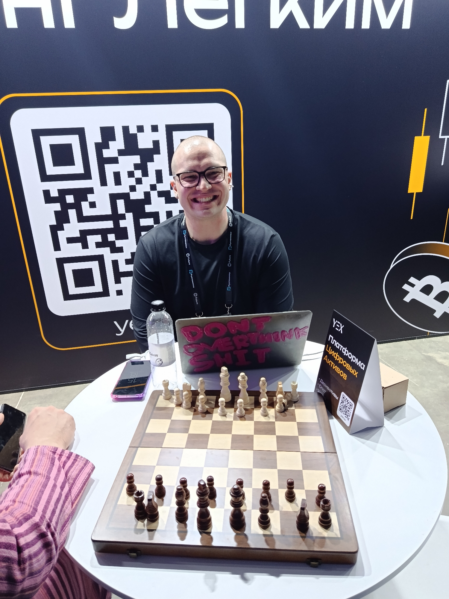 A smiling, bald man with glasses sits behind a table with a chessboard, laptop, and marketing materials. He appears to be at a promotional booth, likely for a cryptocurrency or digital asset platform, as indicated by the "YEX" sign and laptop stickers. The chessboard is set up with pieces arranged in the starting position. A person is partially visible on the left side of the image, holding a smartphone. The booth is against a black background with a large QR code and other graphics. The location seems to be an indoor event, possibly a conference or exhibition.