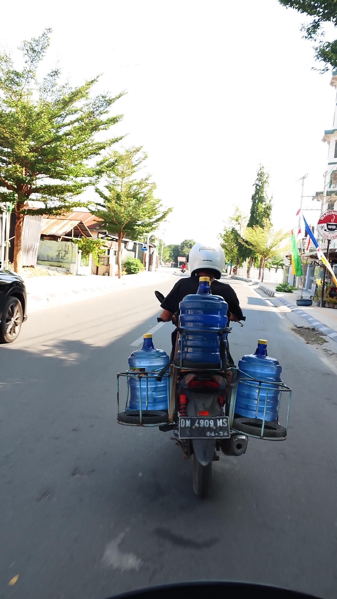 In Balaroa, Indonesia, a person wearing a white HONDA helmet rides a motorcycle carrying five large blue water jugs. The jugs are secured to a metal rack on the back of the motorcycle. The rider, dressed in black, is facing away from the camera. The motorcycle has a black license plate that reads "DN 4909 MS 04-24". The scene takes place on a paved road with trees and buildings lining the sides. A black SUV is partially visible on the left side of the image. The sky is bright and appears to be daytime.