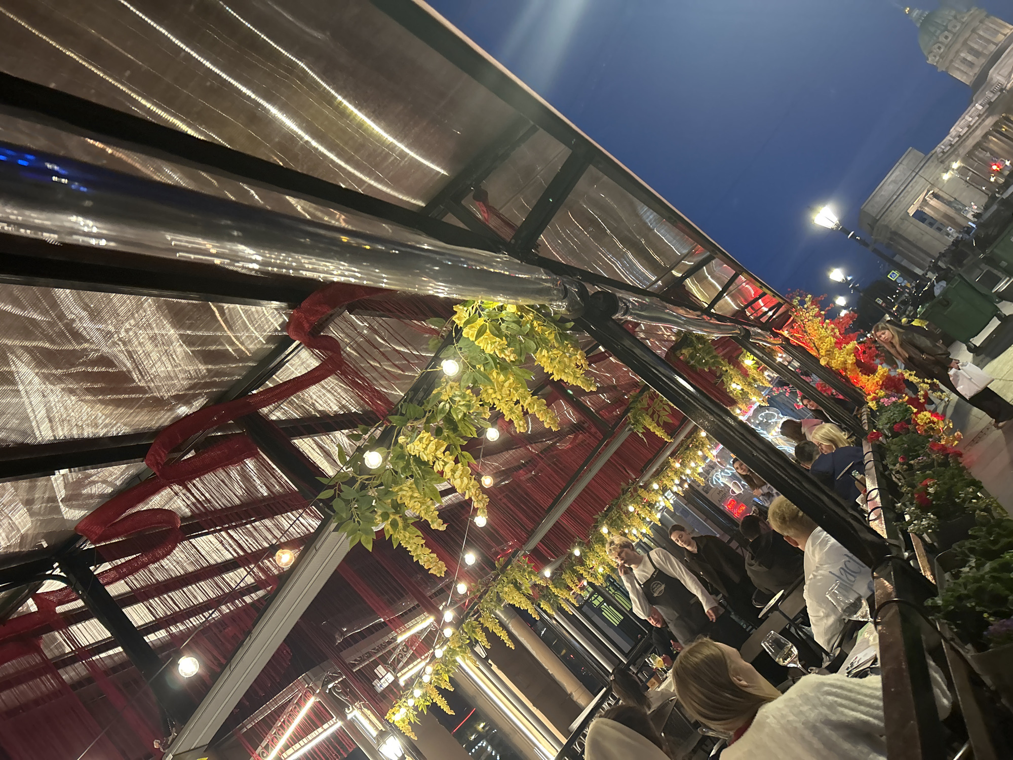 The image depicts an outdoor dining area with a covered roof. Red fabric and yellow floral decorations hang from the roof structure, interspersed with string lights. Several people are seated at tables, some appearing to be dining or socializing. A person in what appears to be a server's uniform is standing and interacting with a table. In the background, a city street is visible with buildings and streetlights, and a large domed building is prominent in the distance, suggesting a significant landmark. The sky is dark blue, indicating it is evening or nighttime.