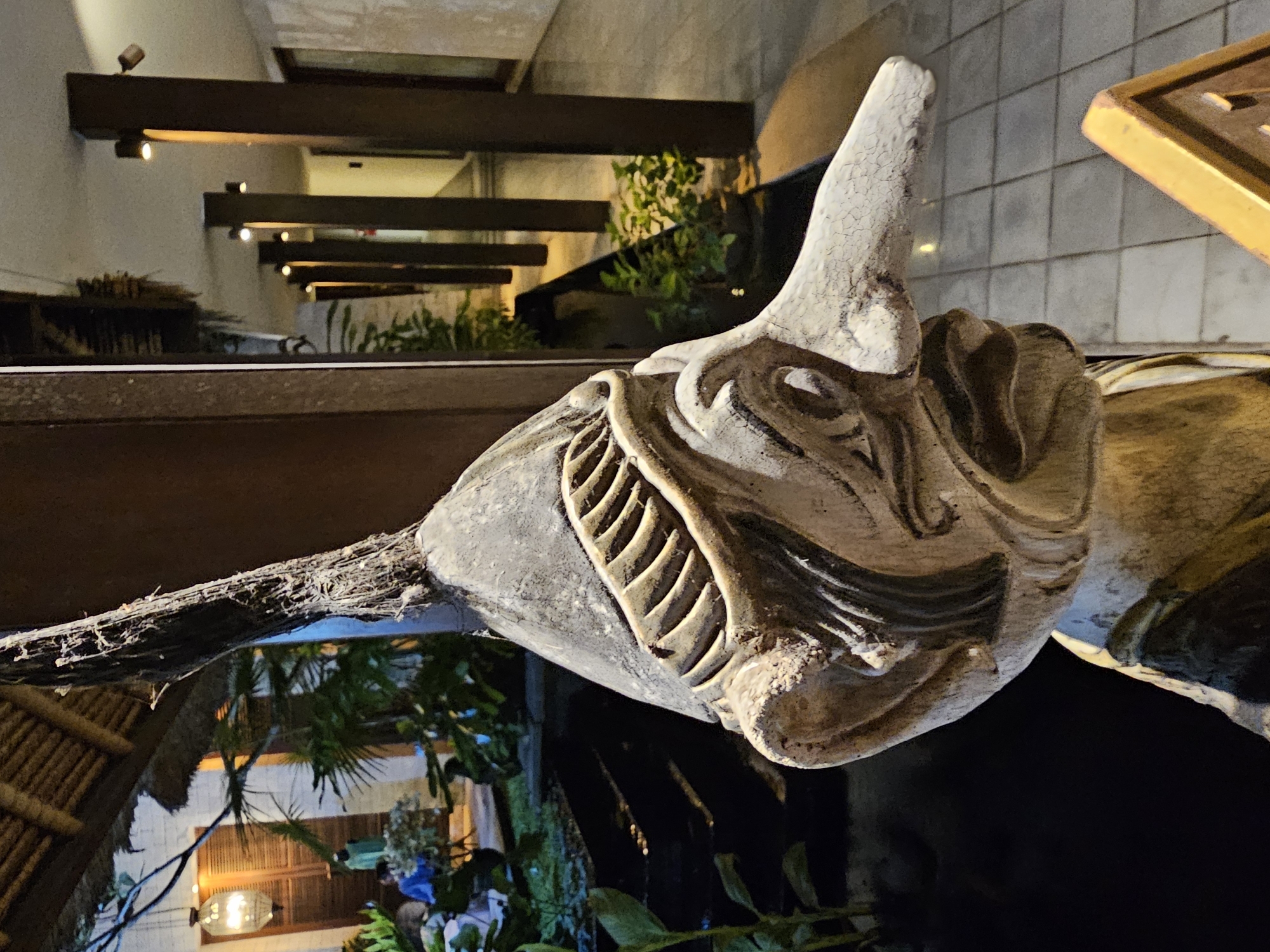 A close-up, rotated image features a carved, weathered stone mask with intricate details and a horn-like protrusion from its forehead. Cobwebs cling to the left side of the mask, adding to its aged appearance. The mask's location is suggested by elements in the background, which show an outdoor setting with tropical plants, water features, and architectural elements such as wooden beams and thatched roofing. The soft lighting and building style suggest it may be in Denpasar, Indonesia.