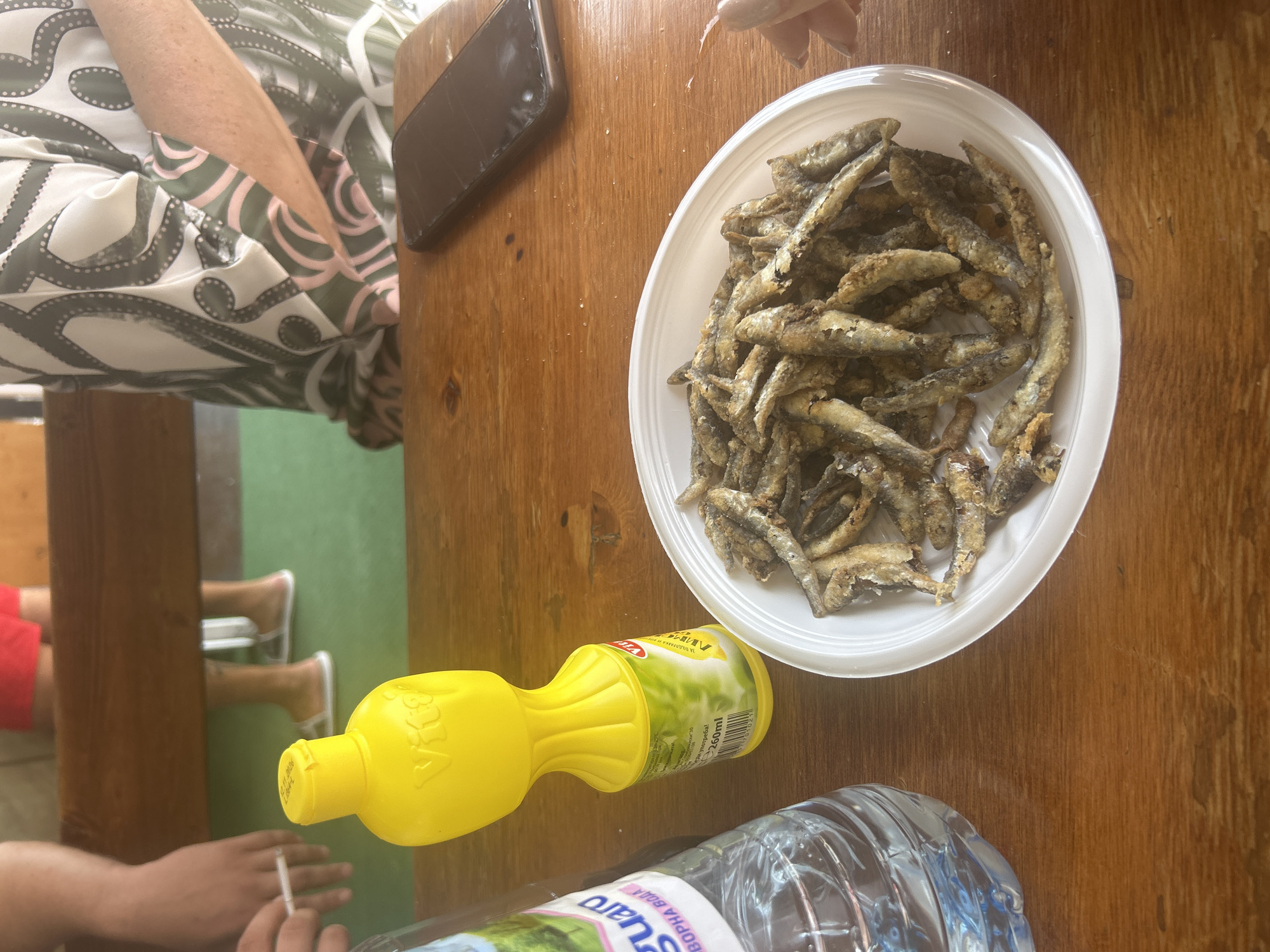 The image shows a close-up of a wooden table with a plate of fried fish, a bottle of water, and a yellow squeeze bottle of lemon juice. In the background, two people are partially visible, one wearing a patterned shirt and the other wearing red shorts. The setting appears to be outdoors, possibly a cafe or restaurant, with a green surface visible behind the table. A smartphone is also present on the table. The fried fish are small and whole, covered in a golden-brown batter. The lemon juice bottle is a bright yellow and has a label with text and an image of a lemon. The water bottle is clear plastic and filled with water.