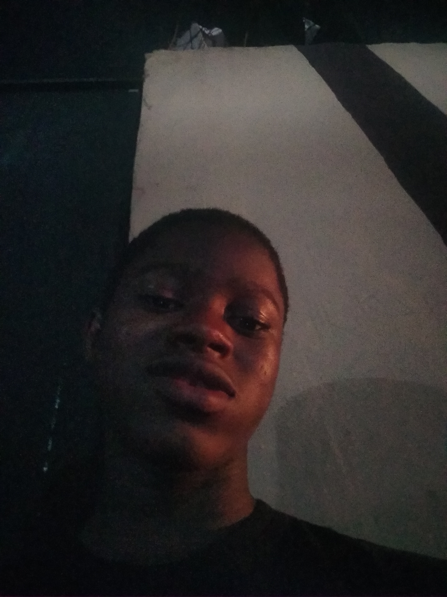 A close-up photo shows a dark-skinned individual in a dimly lit indoor setting. The person is facing the camera with a slight upward angle. Part of a light-colored wall is visible behind the person, with a dark shadow cast on it. There also appears to be a darker object or wall to the left of the person. The location is specified as Shangisha, Nigeria.