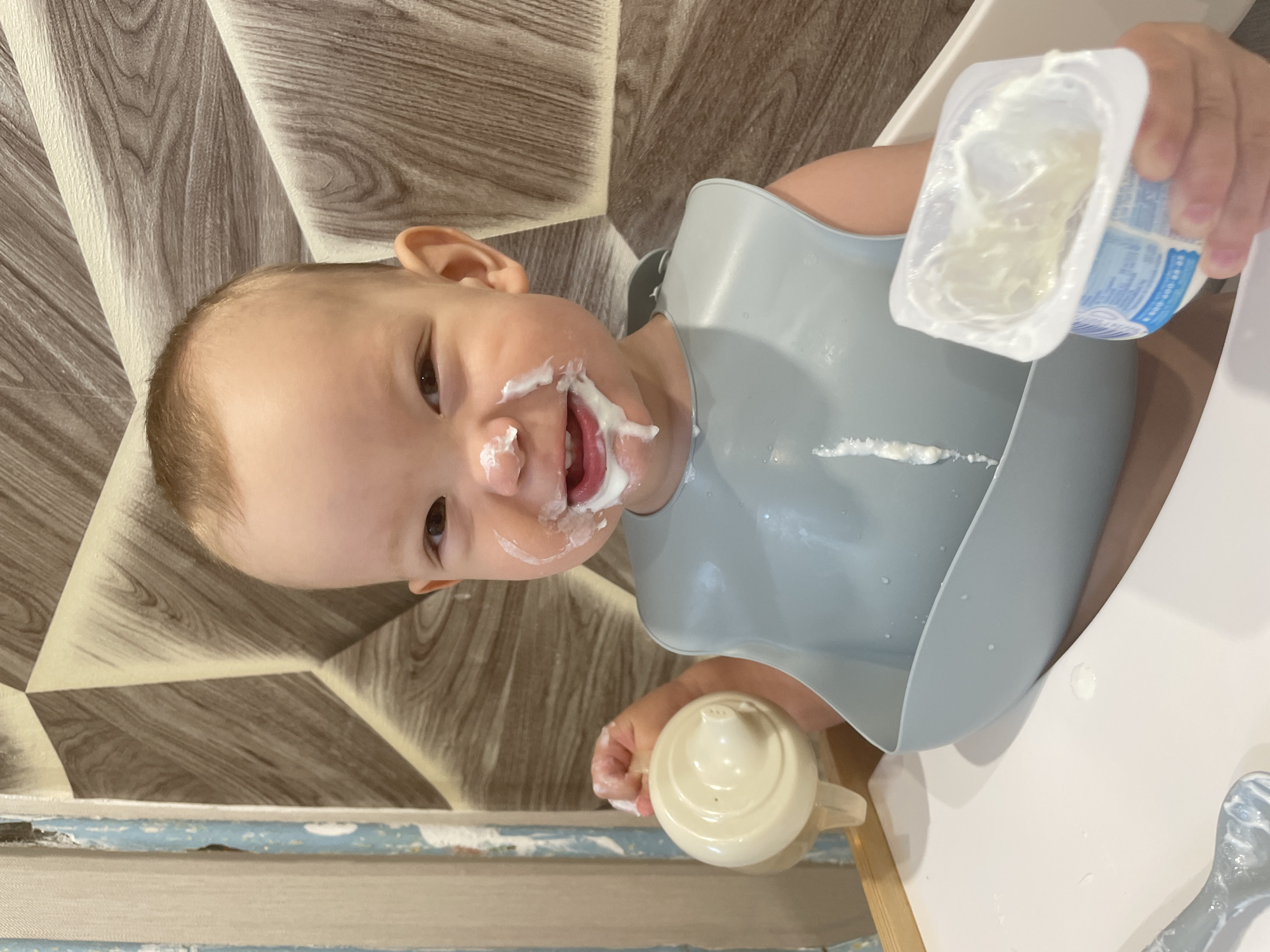 A young child in a high chair is covered in yogurt. The child has yogurt on their face, bib, and hands. The child is holding a sippy cup in one hand and a container of yogurt in the other. The yogurt is smeared on the tray of the high chair. The background is a wall with a geometric pattern. The child is located in Perm, Russia.