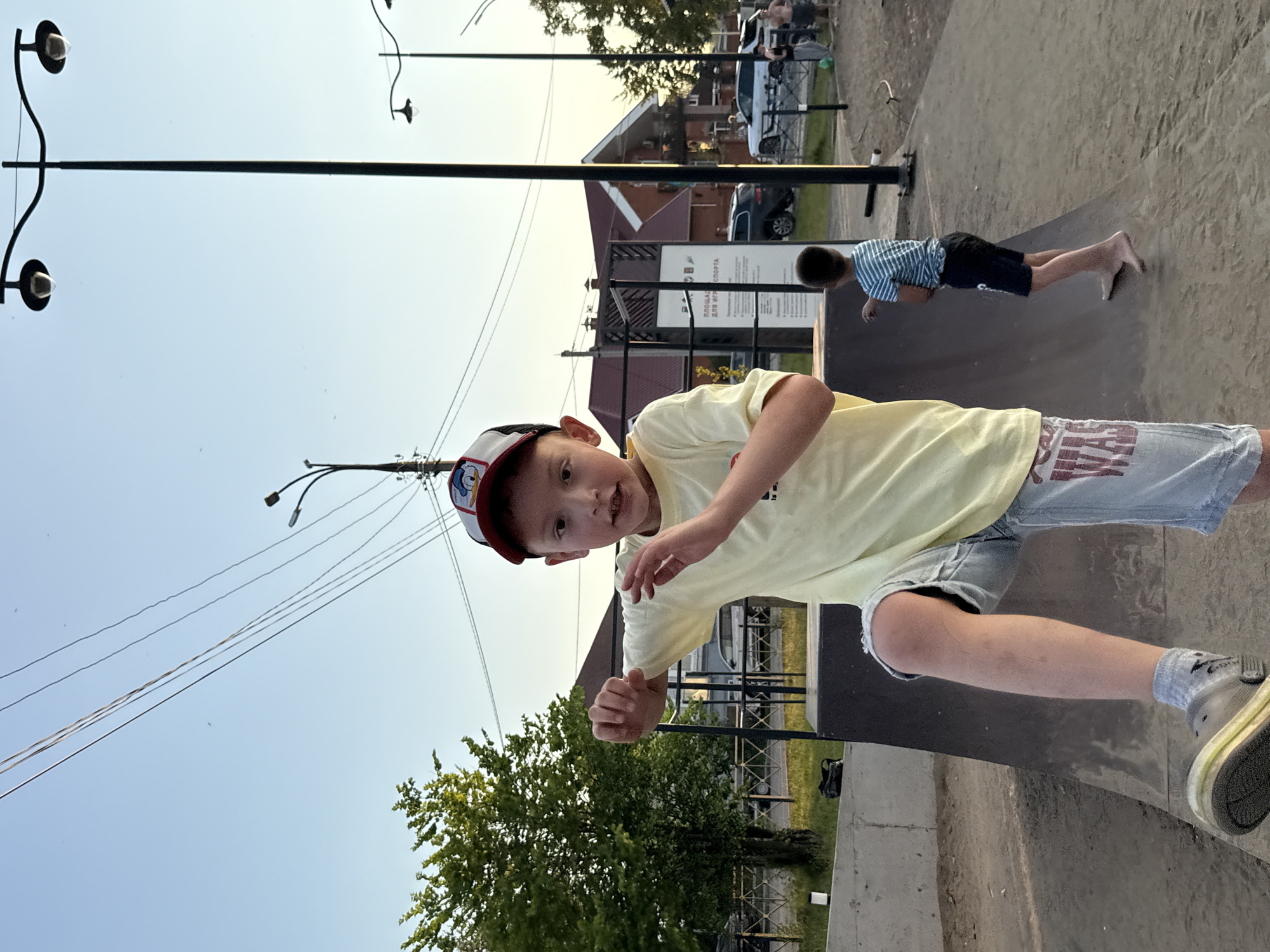 In Votkinsk, Russia, at an outdoor sports park, a young boy wearing a yellow t-shirt and denim shorts stands on a ramp. He has a baseball cap on his head and is in motion, with his right arm extended. Behind him, another boy in a striped t-shirt and black shorts stands on the ground, barefoot. The park features ramps and other sports equipment, with trees and buildings visible in the background under a clear sky. Streetlights are present, and several cars and people are in the distance.