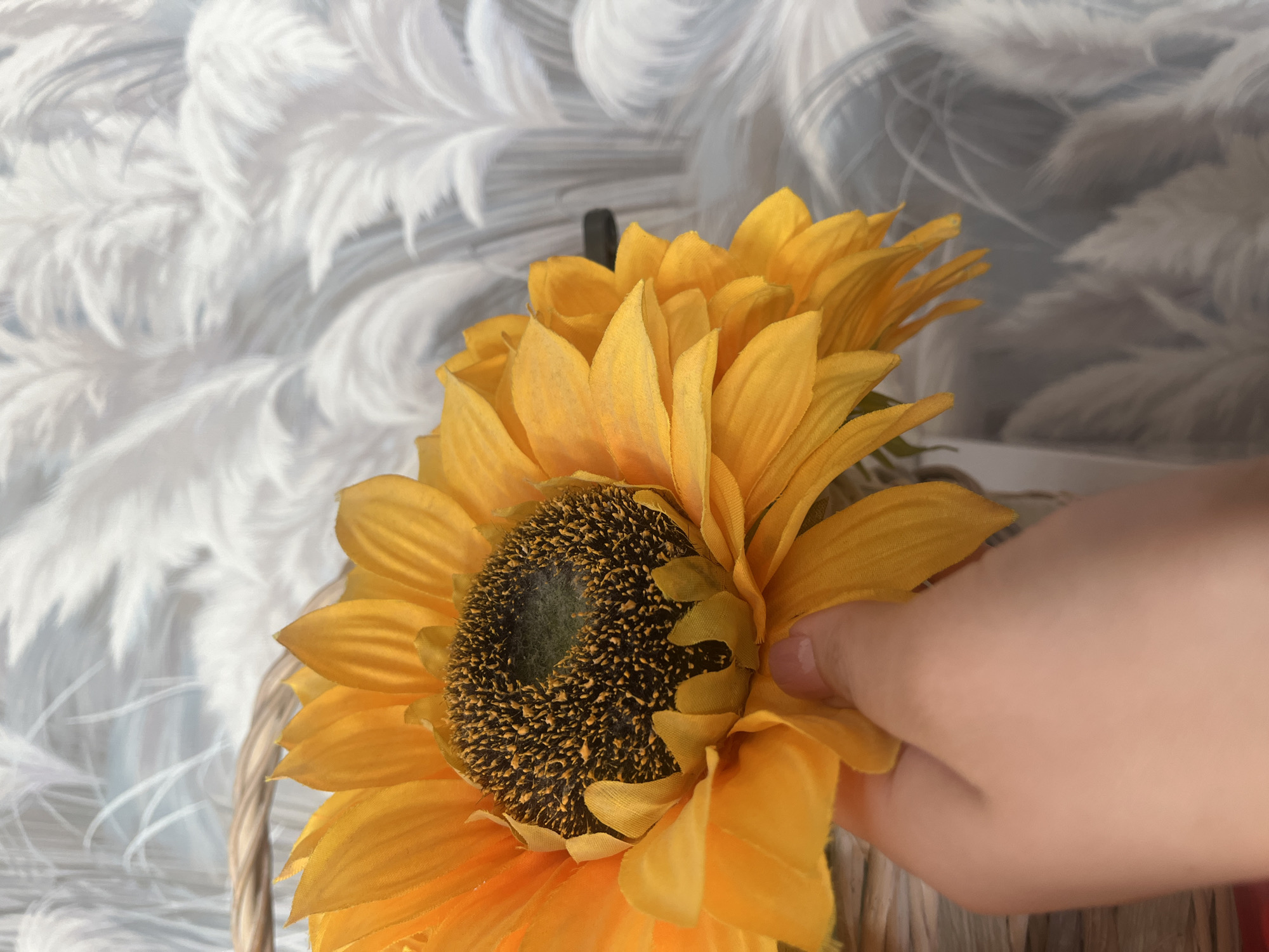 A person's hand, with light skin and short fingernails, is holding the yellow petals of a large artificial sunflower. The sunflower is in a woven basket. The background is a wall with a pattern of feathery white plumes against a greyish-blue backdrop. There is no indication of the location being in Бруснятское, Russia; this image depicts an indoor scene.