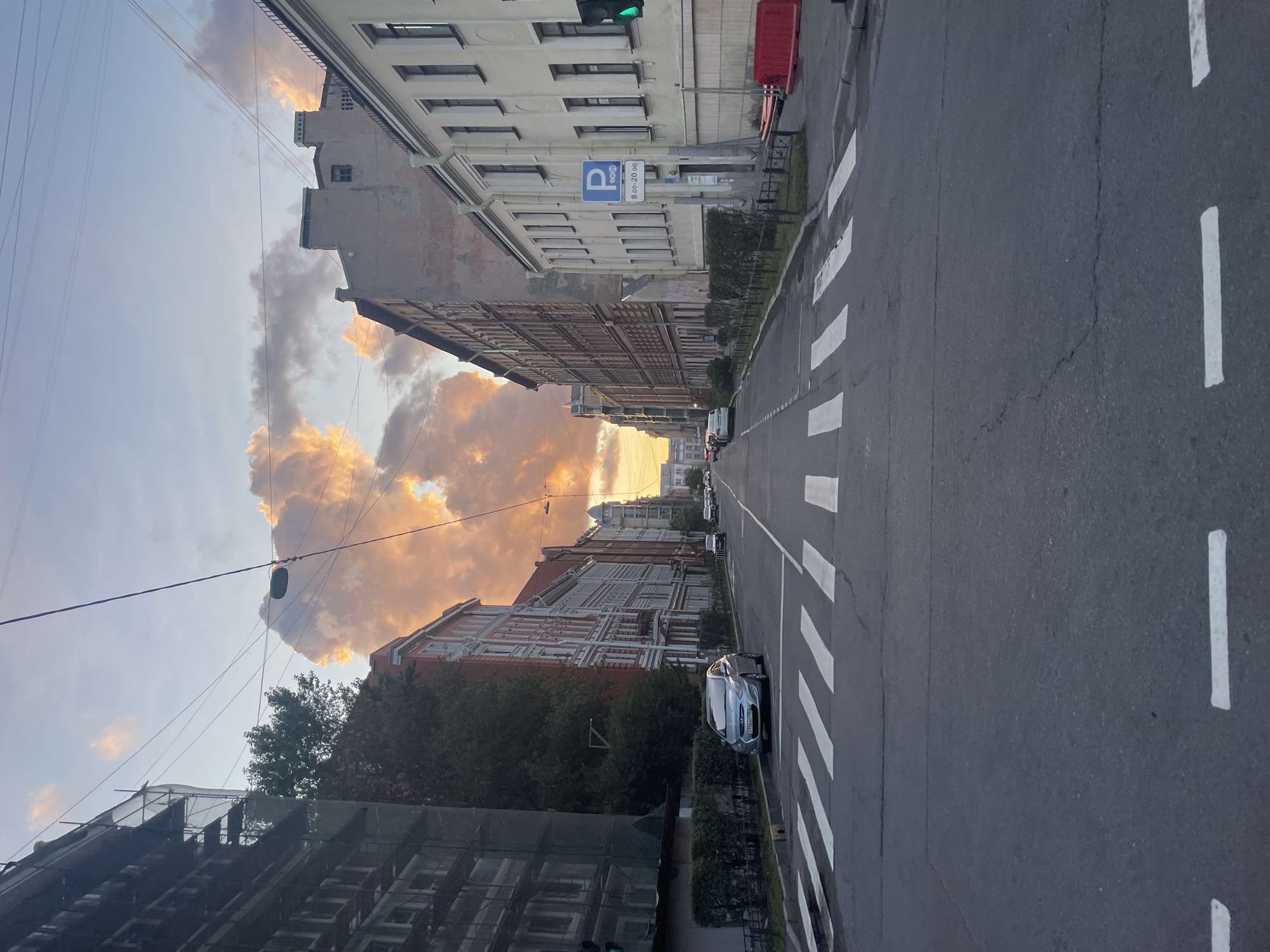 The image shows a street in Saint Petersburg, Russia, during sunset. The sky is filled with dramatic, orange-tinged clouds. The street is paved and has white pedestrian crossings. On either side of the street are multi-story buildings with classic European architecture. Some buildings appear to be under renovation, with scaffolding visible. There are several cars parked along the street, and some are driving in the distance. A traffic light is visible on the right side of the image, showing a green signal. A parking sign is also visible. The overall atmosphere is calm and serene, with the warm colors of the sunset creating a beautiful backdrop.