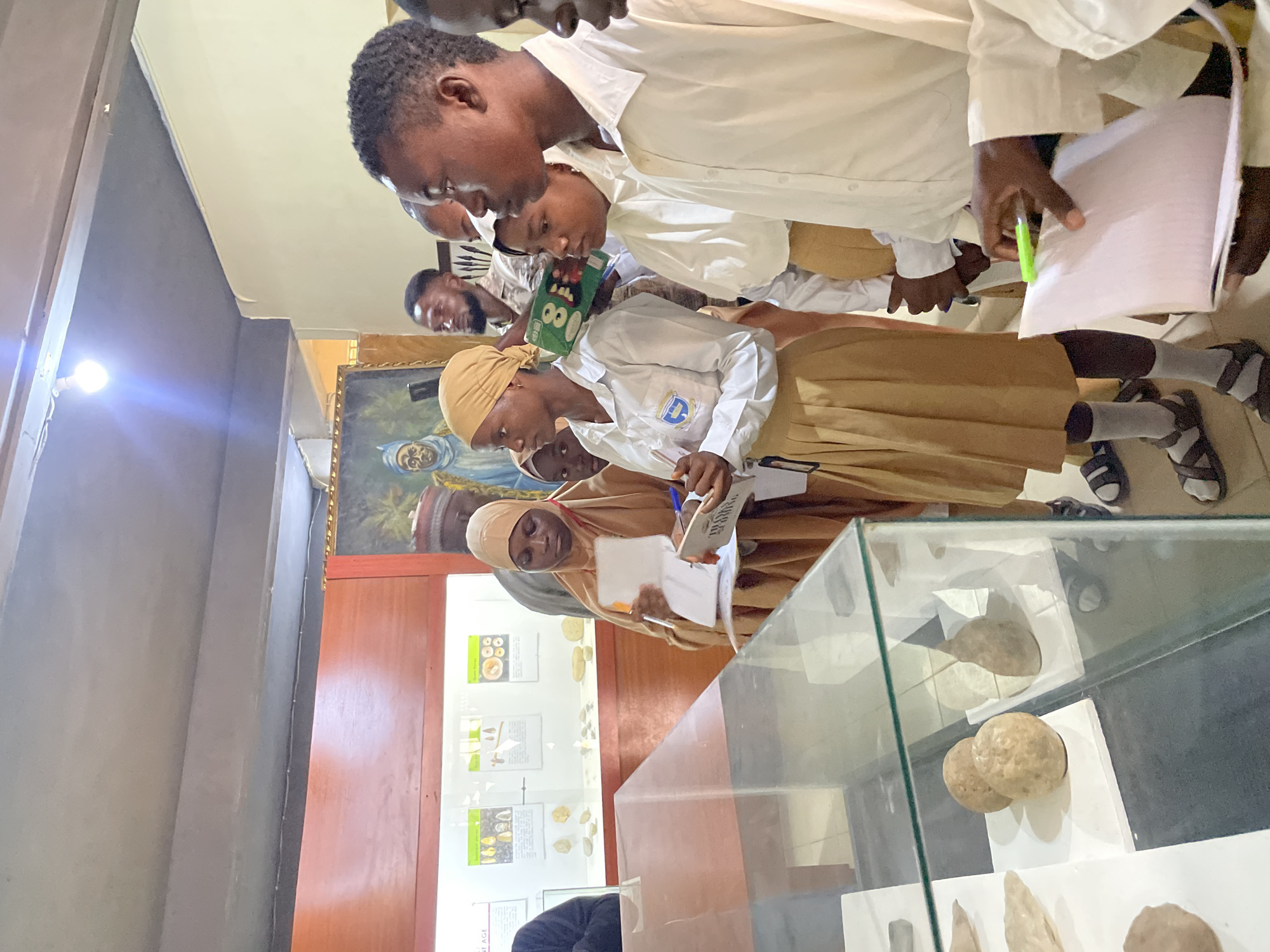 The image shows a group of young people, likely students, observing exhibits in a museum or historical display. The subjects are predominantly of Nigerian descent, dressed in school uniforms. Several individuals are seen taking notes or looking intently at the displays, suggesting an educational visit.
The scene is set indoors, with a museum-like environment. In the background, a large framed painting depicts a figure, possibly historical or religious. Display cases with artifacts are visible, featuring what appear to be geological or archaeological specimens. One display case prominently shows rounded stones or rock samples. Educational posters with text and images are mounted on the walls. The lighting suggests it is daytime, though no specific weather conditions can be inferred.
The activity appears to be a guided tour or a field trip, where students are engaged in learning and documentation. The students' focus and note-taking indicate a serious approach to the educational experience. Their attire, consisting of white shirts and tan skirts or trousers, points to a formal school setting. The overall atmosphere seems calm and studious.
Notable details include the cultural context suggested by the clothing and the painting. The presence of display cases and informative posters confirms the setting as an educational institution or a public display of historical or cultural artifacts. No specific time of day or weather information is discernible. The emotions conveyed are primarily concentration and curiosity. There is some visible text on the educational posters, but it is not legible enough to be transcribed.
