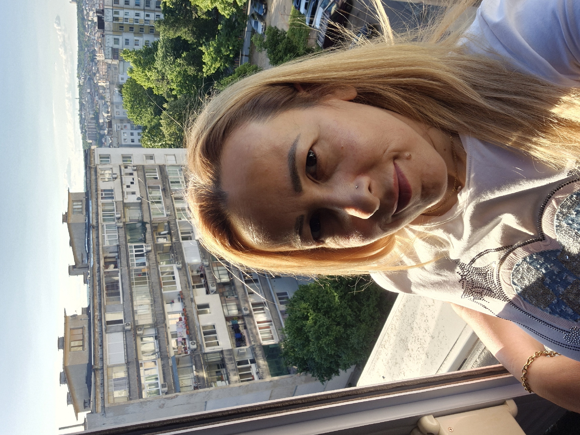 A woman with blonde hair and a nose piercing smiles at the camera in front of an apartment building. She is wearing a white t-shirt with a sparkly design. The building appears to be in a city with other buildings and trees visible in the background. The sky is clear and blue, suggesting a sunny day. The image is taken from a slightly high angle, possibly from a balcony or window.