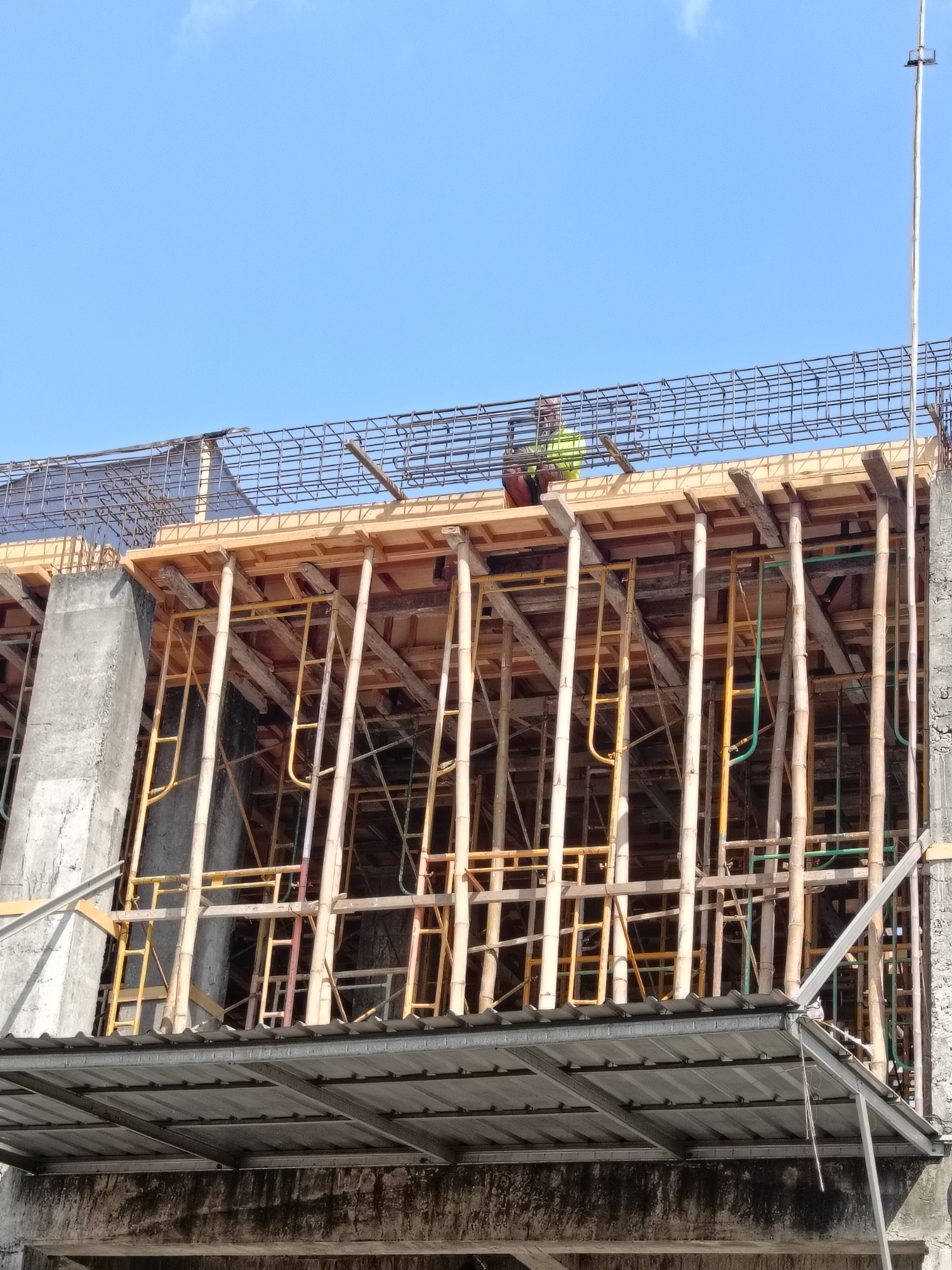 A low-angle, medium shot captures a construction site with a bright blue sky and a few wispy clouds in the background. In the foreground, a metal roof extends from the bottom of the frame upwards. Above this, a complex structure of scaffolding, made of both bamboo and yellow metal poles, supports a wooden platform. Several wooden beams are also visible, forming part of the construction. On the top level of the scaffolding, a construction worker wearing a bright yellow vest and a hard hat is partially visible, appearing to be working on the metal reinforcement bars that are being laid out. To the left of the scaffolding, a large, rough concrete pillar rises towards the sky. The overall scene depicts an active construction project, likely for a building, in Gedangan, Indonesia.