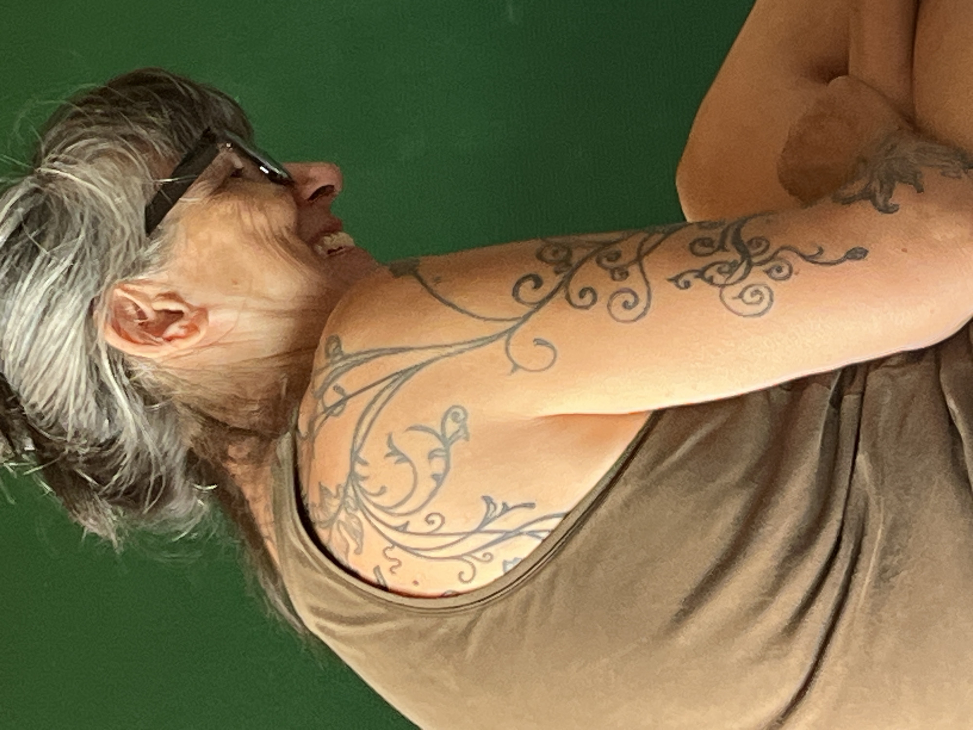 A close-up profile shot captures a woman with grey hair pulled back and wearing glasses, laughing with her head tilted down and to the side. She is wearing a tan tank top and has a tattoo of swirling vine-like patterns on her left shoulder and arm. The background is a solid green color. The image was taken in Palavas-les-Flots, France.
