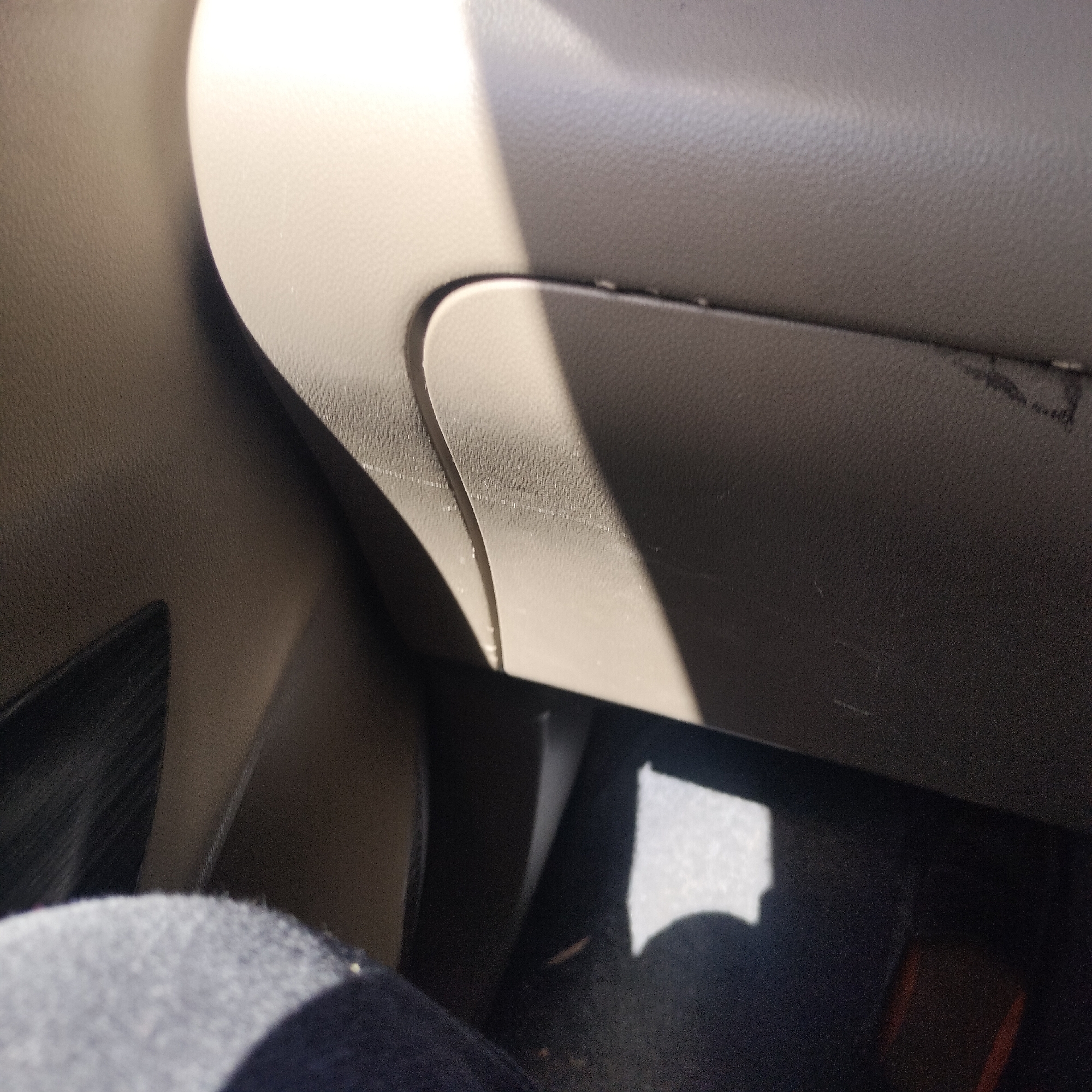 The media file captures the interior of a car, likely from the perspective of a passenger. The central subject is a light beige or tan-colored glove compartment door on the dashboard, featuring a distinct, slightly curved line that suggests its opening mechanism or a design element. Bright sunlight enters the vehicle from the upper right, creating a prominent diagonal highlight across the glove compartment's surface and casting sharp shadows. Faint scratch marks are discernible on the glove compartment.
To the left of the glove compartment, another textured plastic panel, in a darker shade, is visible. The overall color palette of the car interior consists of light beige/tan and darker grey/black tones. In the bottom left corner, a knee or upper leg, covered in dark fabric, is partially visible, indicating a person is seated in the vehicle. The floor area is dark, possibly a car mat or carpet, with a patch of light towards the bottom right.
The scene suggests a sunny daytime setting. No specific activity is taking place beyond being inside a stationary or moving car. There is no visible text in the image. The location context provided is Tanjongkeumala, Indonesia.