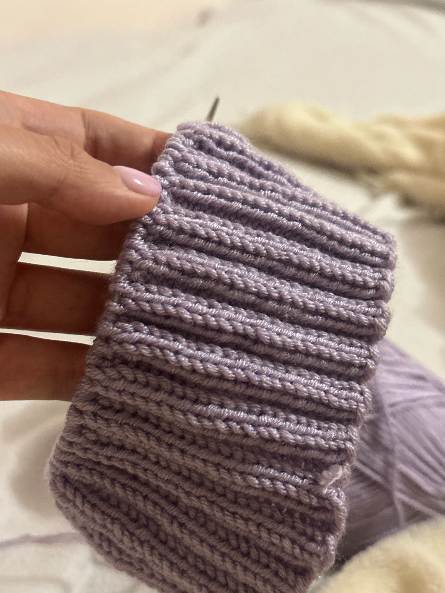 A person's hand with light pink nail polish is holding a piece of knitted fabric in a ribbed pattern. The fabric is a light purple color. A knitting needle is visible in the upper right portion of the fabric. In the background, there is a ball of light purple yarn and a blurry, light-colored fuzzy object. The scene appears to be indoors on a white surface, possibly a bed or a table covered with a white sheet. There is no indication of the location being in Gyumri, Armenia.