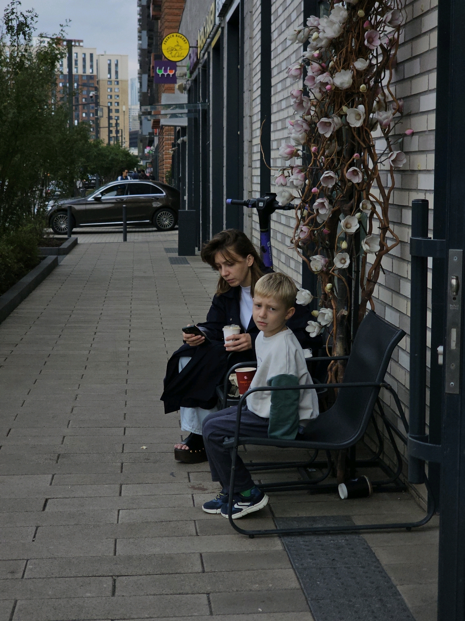 A woman and a young boy are sitting on a bench outside a building in Moscow, Russia. The woman, on the left, is wearing a dark jacket and light blue jeans. She is holding a mobile phone in her right hand and a paper cup in her left. The boy, on the right, is wearing a light-colored sweatshirt and dark pants. He is looking at the camera. A decorative tree with artificial flowers is behind them, and a brick wall forms the background. A dark-colored car is parked on the street to the left of the scene. The ground is paved with rectangular blocks.