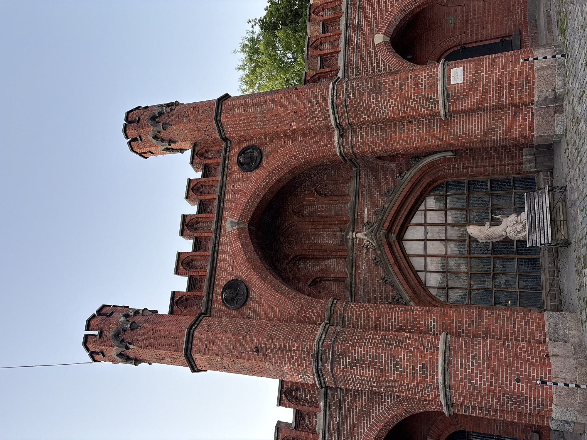 The image shows a detailed view of the Brandenburger Tor in Kaliningrad, Russia. The iconic red brick structure features two prominent towers with crenellated battlements and arched windows. A large, arched stained-glass window is visible in the center, adorned with decorative stonework above. To the left of this window, a stone sculpture of a fish is placed on a bench, with a tree visible in the background on the right. The foreground is paved with cobblestones. The sky is clear and blue.