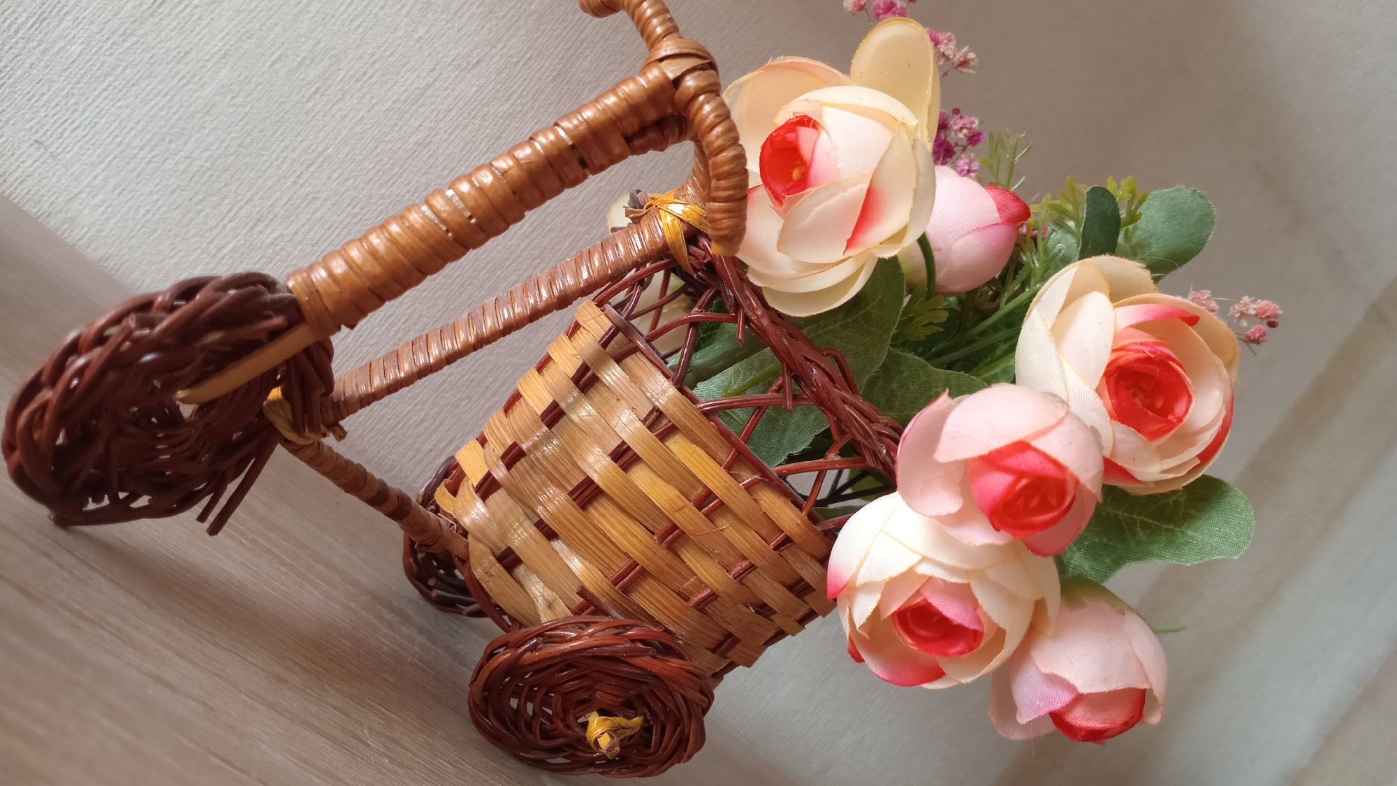 A close-up, angled shot shows a decorative wicker bicycle with a basket filled with artificial flowers. The bicycle is made of light brown and dark brown wicker, with a woven basket attached to the front. The flowers are a mix of light pink, cream, and red, with green leaves. The background is a textured light gray wall. The scene is indoors and does not contain any people or actions.