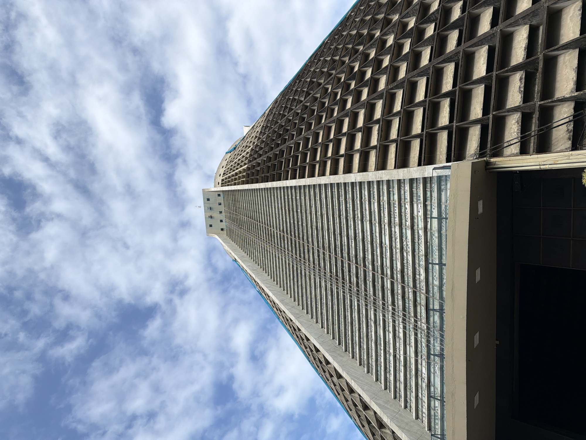 The image shows a low-angle view of the Metropolitan Cathedral of Saint Sebastian in Rio de Janeiro, Brazil. The cathedral, a modern architectural landmark, is characterized by its conical shape and stained-glass windows. The exterior is made of concrete with a grid-like pattern. The sky above is a mix of blue and white clouds. The perspective emphasizes the height and unique design of the building, drawing the viewer's eye upwards towards the apex.