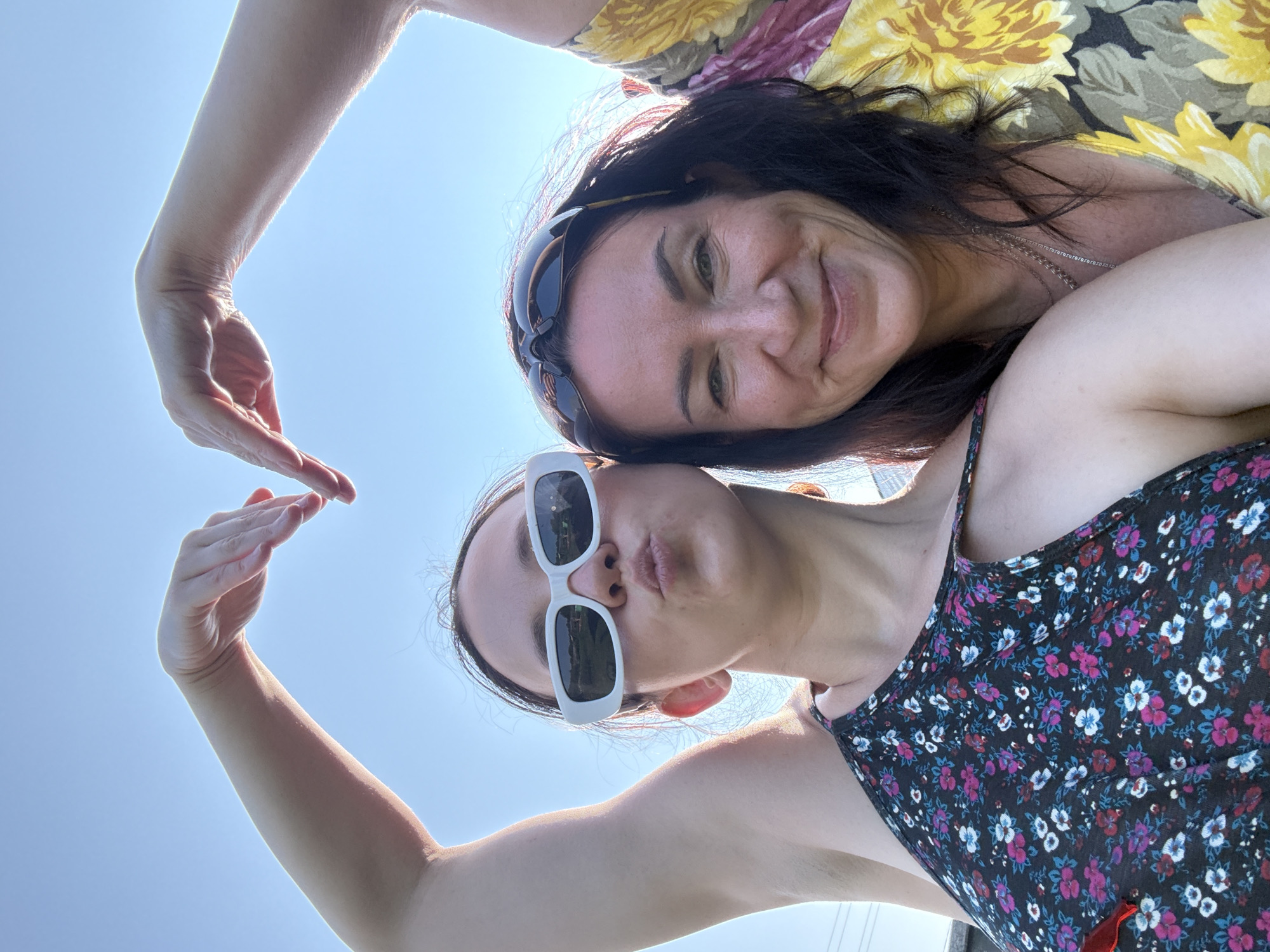 Two women form a heart shape with their arms against a bright blue sky. The woman on the left, wearing white rectangular sunglasses and a floral-print halter top, puckers her lips. The woman on the right, wearing dark sunglasses on top of her head and a yellow floral-print top, smiles at the camera. Both women appear to be outdoors, with the sky being the dominant background element. There is no information about the city of Zasechnyi, Russia, visible in the image.