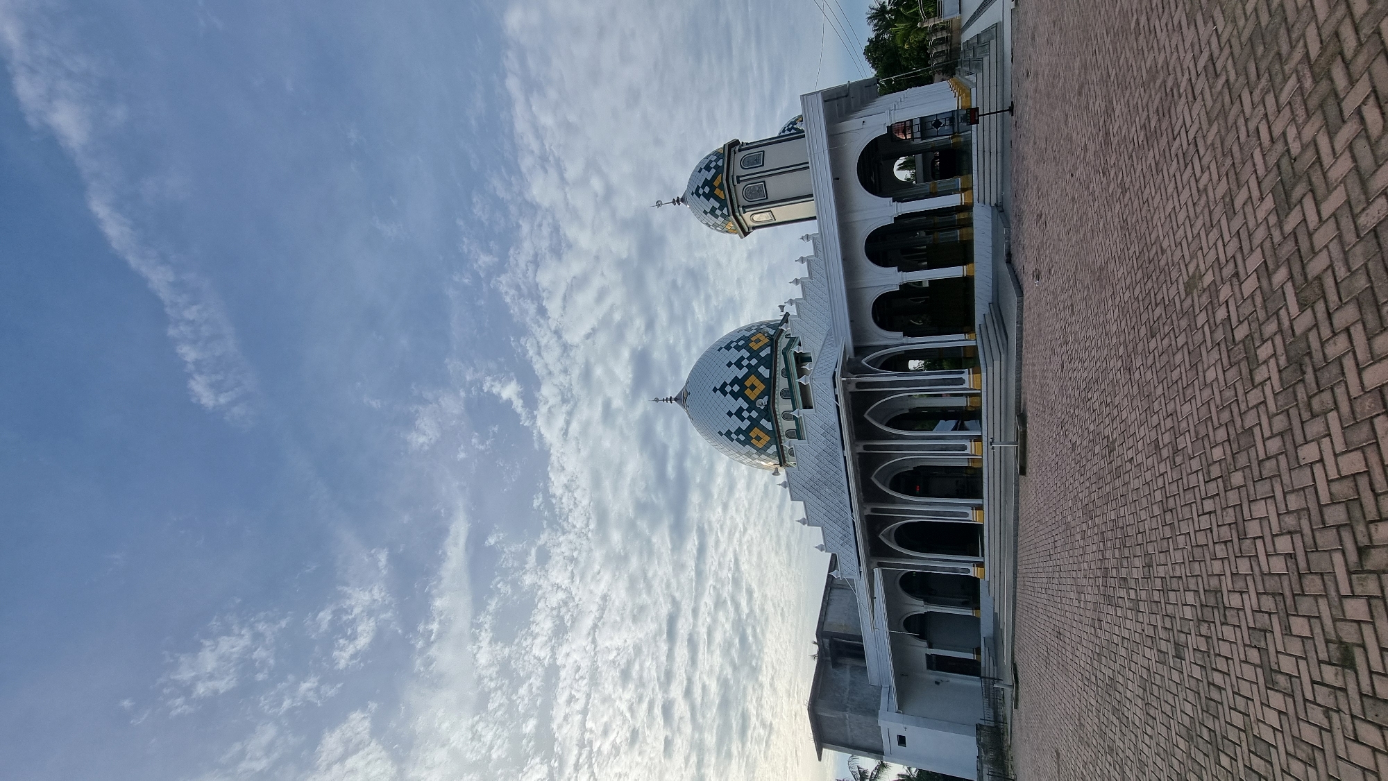 The image depicts a mosque in Meunasahlhok, Indonesia, with a cobblestone courtyard in the foreground. The mosque has a white facade with arched walkways supported by pillars. It features a prominent central dome with a decorative pattern of blue, white, and yellow diamonds, and a smaller dome on a separate tower. The sky above is a clear blue with scattered white, wispy clouds, suggesting it might be early morning or late afternoon. The surrounding environment appears to be a mix of urban and tropical, with hints of trees visible to the right of the mosque. There are no people or animals visible in the scene.