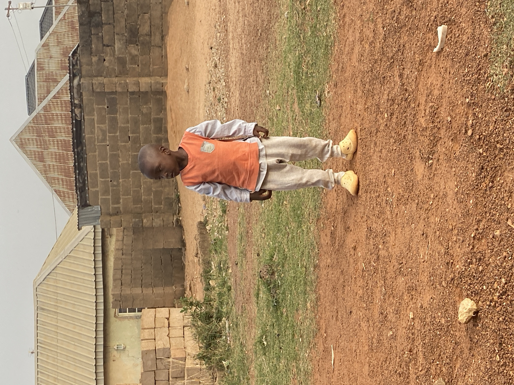 The image, captured in Bukuru, Nigeria, features a young child standing in an outdoor, unpaved area. The child, likely a boy, has short dark hair and is dressed in an orange sleeveless vest worn over a light-colored, long-sleeved shirt. He wears light-toned trousers and bright yellow Crocs-style footwear. He is standing still, looking downwards and slightly to his left.
The background shows a simple, somewhat undeveloped setting with two buildings. One building, more prominent, has a rusty corrugated metal roof and walls constructed from concrete blocks. Another building, further left, features a lighter, possibly silver, corrugated metal roof. Building blocks are stacked near the base of one structure, which also includes a small window.
The ground consists primarily of reddish-brown earth and small gravel, interspersed with patches of green grass. Various small rocks and debris are scattered across the ground, including a distinctive white, irregular object located to the child's right. A small green bush is visible in the lower left section of the frame.
The scene is lit by soft, diffused daylight, suggesting either an overcast sky or a time of day without harsh sunlight. The child is not engaged in any specific activity, simply standing. No visible text is present in the image.
