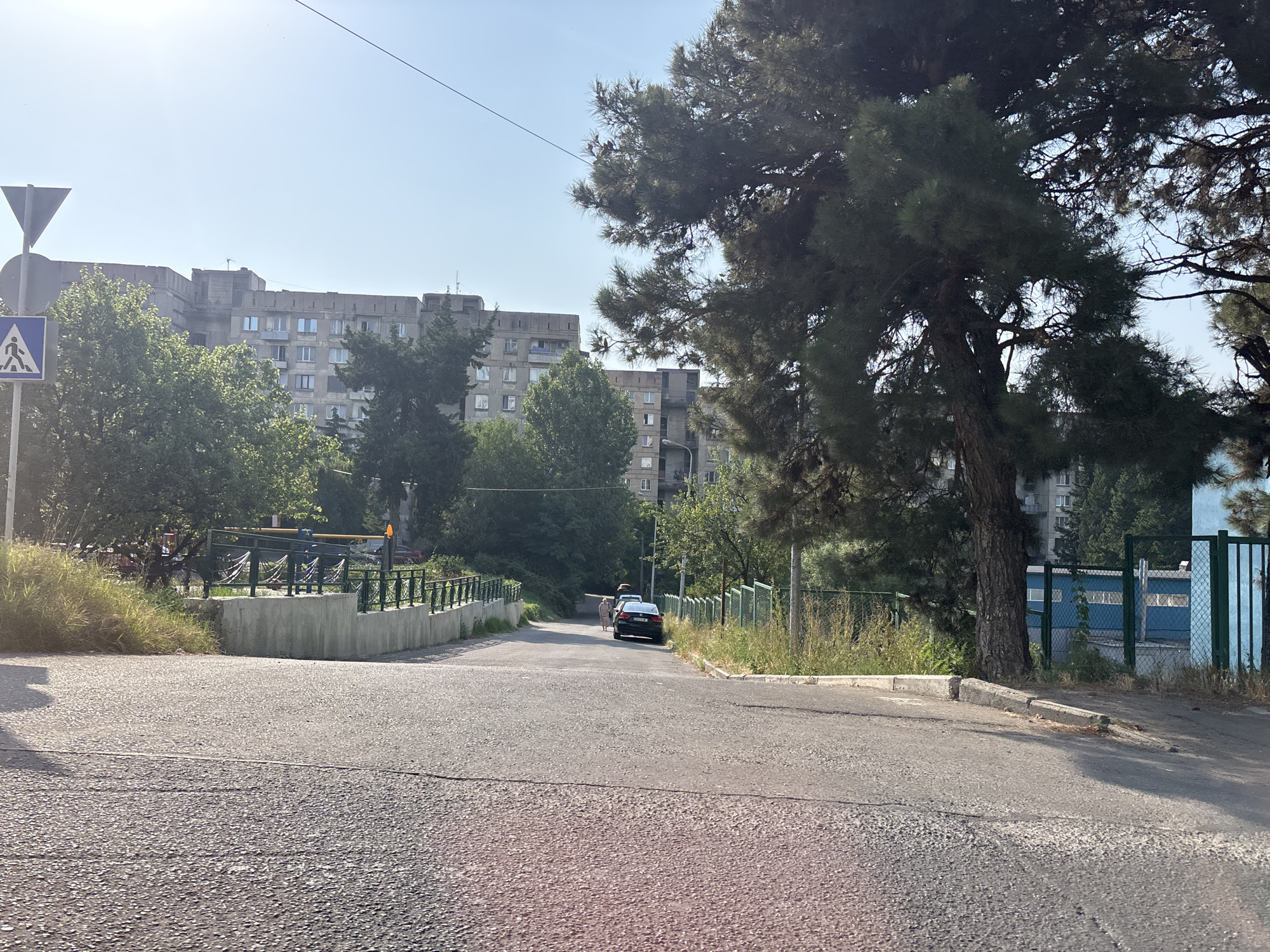 The image shows a street in Tbilisi, Georgia, on a sunny day. In the foreground, the asphalt road curves slightly upwards and to the right. On the left side of the road, there is a concrete barrier topped with a metal railing. Beyond the barrier, there is overgrown grass and trees. In the distance, two cars are parked on the side of the road, and a person is walking away from the camera. Behind the road, there are several large, multi-story apartment buildings with a Brutalist architectural style. Tall trees are scattered throughout the scene, some with dense foliage. A blue and green fence runs along the right side of the road. A pedestrian crossing sign is visible on the left side of the image, near the beginning of the road. The sky is clear and bright blue.