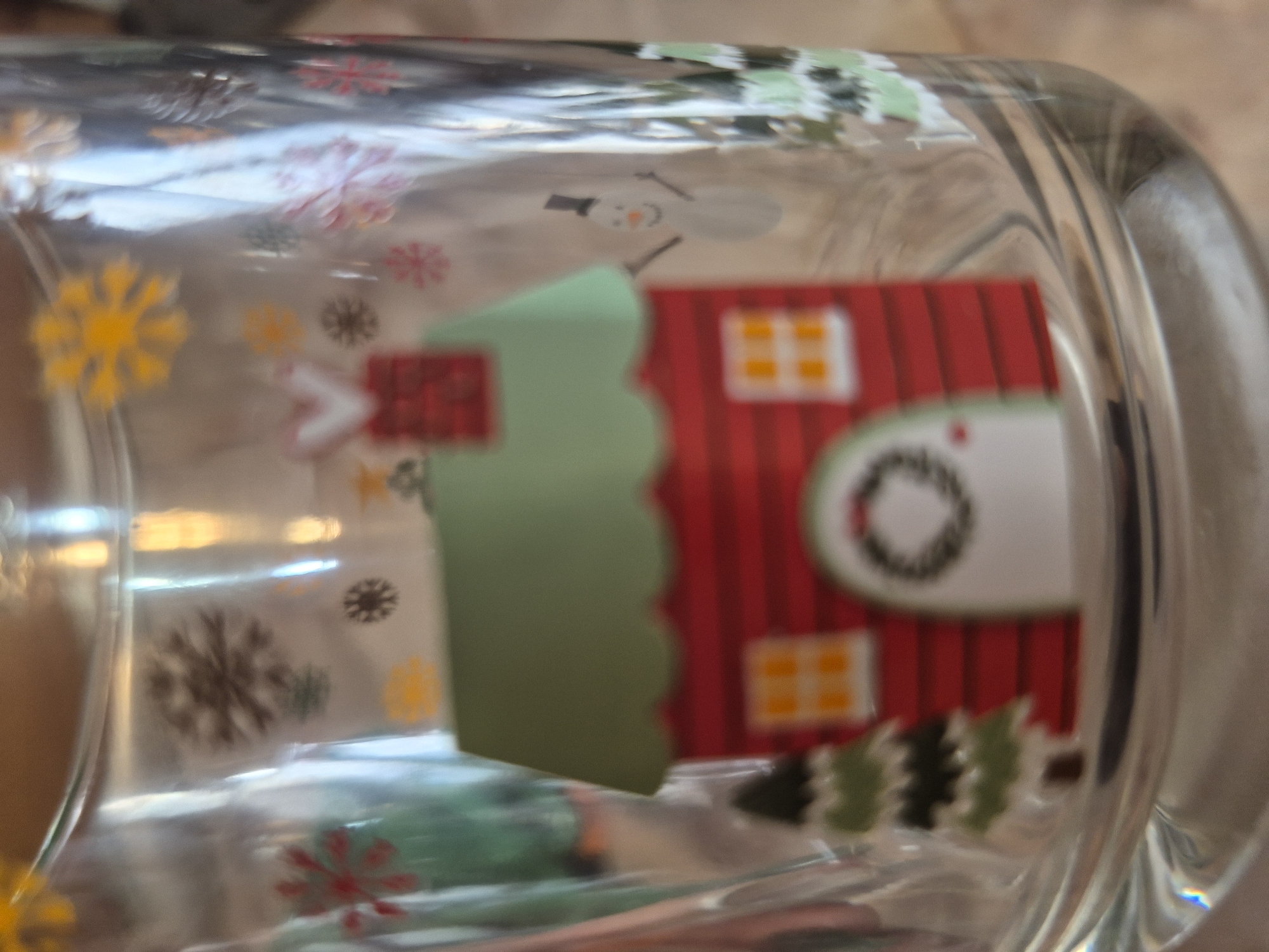 The primary subject of the image is a clear glass drinking vessel, possibly a mug or tumbler, adorned with a festive, winter-themed design. The glass is depicted lying on its side, showcasing its decorated surface.
The design prominently features a stylized red house with vertical stripes, a light green scalloped roof, and a small red chimney. Two square orange windows with white outlines are visible on the right side of the house, and a white circular wreath with a dark outline is placed on its front. To the left of the house, a white snowman is illustrated, complete with a black top hat, black stick arms, and an orange carrot nose. Below the house, several small evergreen trees in shades of green and black add to the scene.
Numerous snowflakes of various sizes and colors, including bright yellow, dark brown/black, red/pink, and light green, are scattered across the glass, enhancing the winter holiday atmosphere. The image is a close-up shot, and some areas are slightly out of focus, though the main design elements are clearly discernible.
The scene depicted on the glass is a cheerful winter landscape. The object itself appears to be indoors, placed on a softly blurred light brown or beige surface, possibly a wooden table or countertop. No specific activity is taking place; the image is a still life. The designs evoke a sense of warmth and holiday spirit, characteristic of the winter season. There is no visible text on the object. The location context for the item is Yerevan, Armenia, though no specific visual cues within the image confirm this.